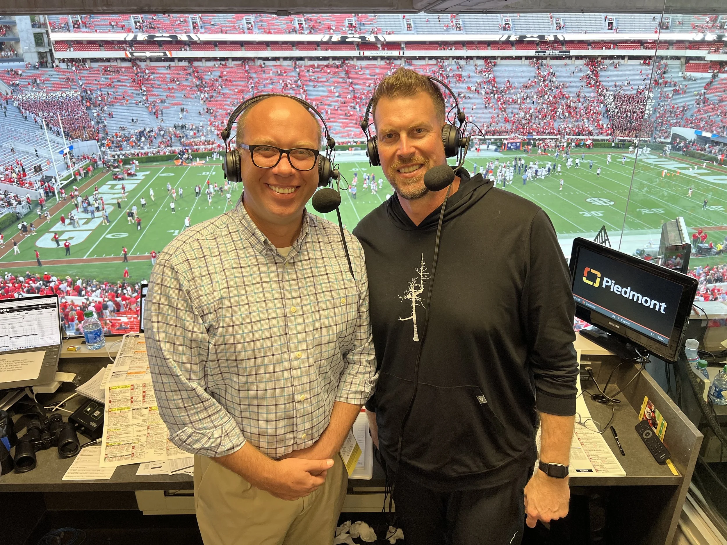Two men in broadcast headsets standing in a sports broadcast booth overlooking a football stadium filled with fans and players.