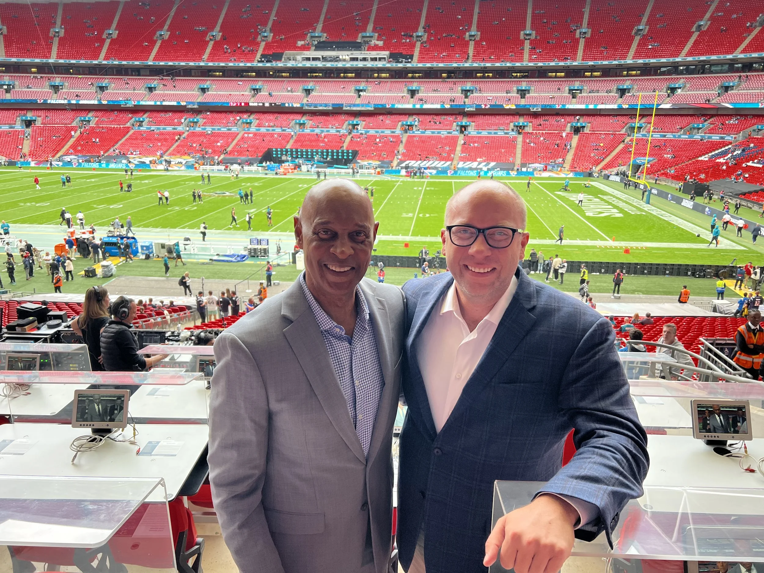 Two men in suits smiling at the camera in a stadium suite overlooking a football field with players and staff preparing on the field.