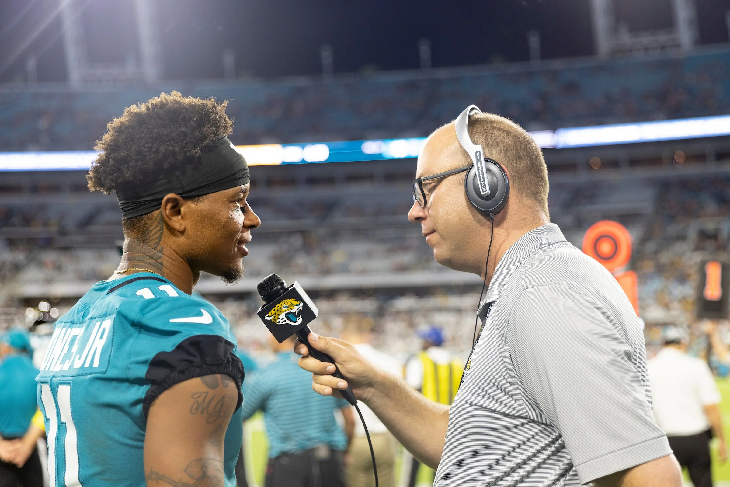 A football player in a teal Jacksonville Jaguars jersey being interviewed by a reporter with headphones on at a stadium.