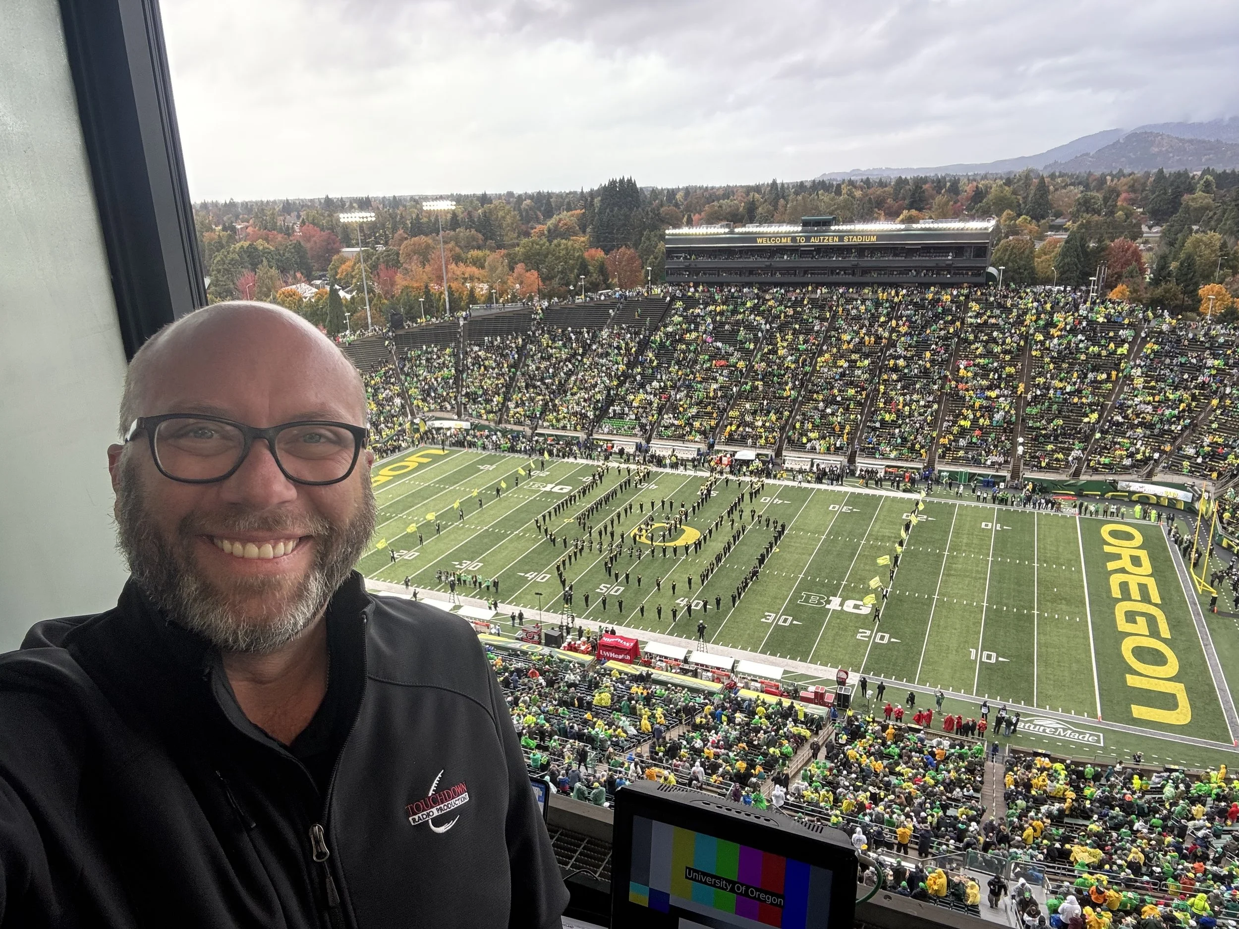 A smiling man wearing glasses and a black jacket inside a stadium box seat, overlooking a football field at Autzen Stadium in Oregon, with fans in the stands and a marching band on the field, during a game with fall foliage and mountains in the backg