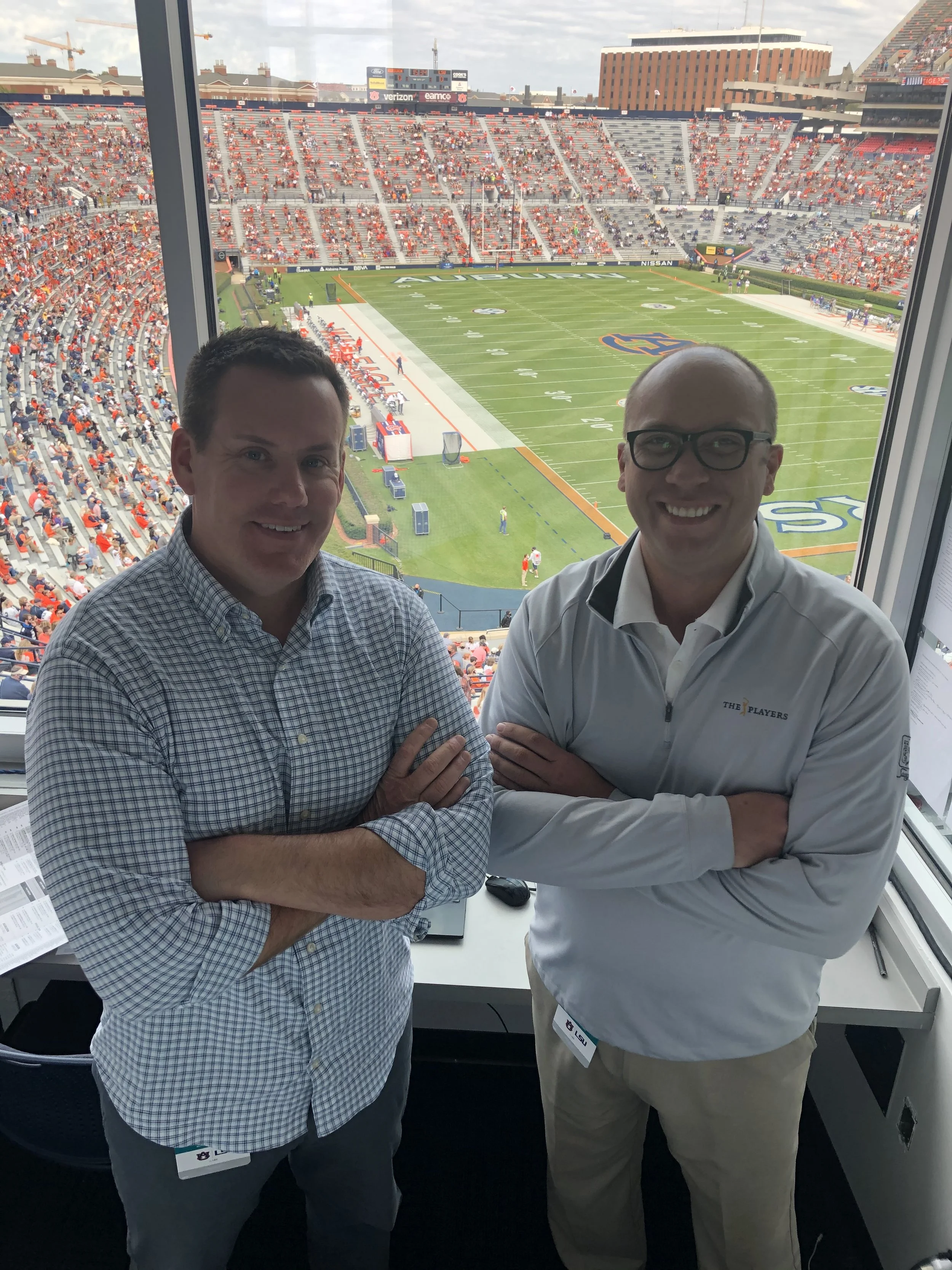 Two men smiling with crossed arms inside a press box at a football stadium, with the field and spectators visible through large windows behind them.