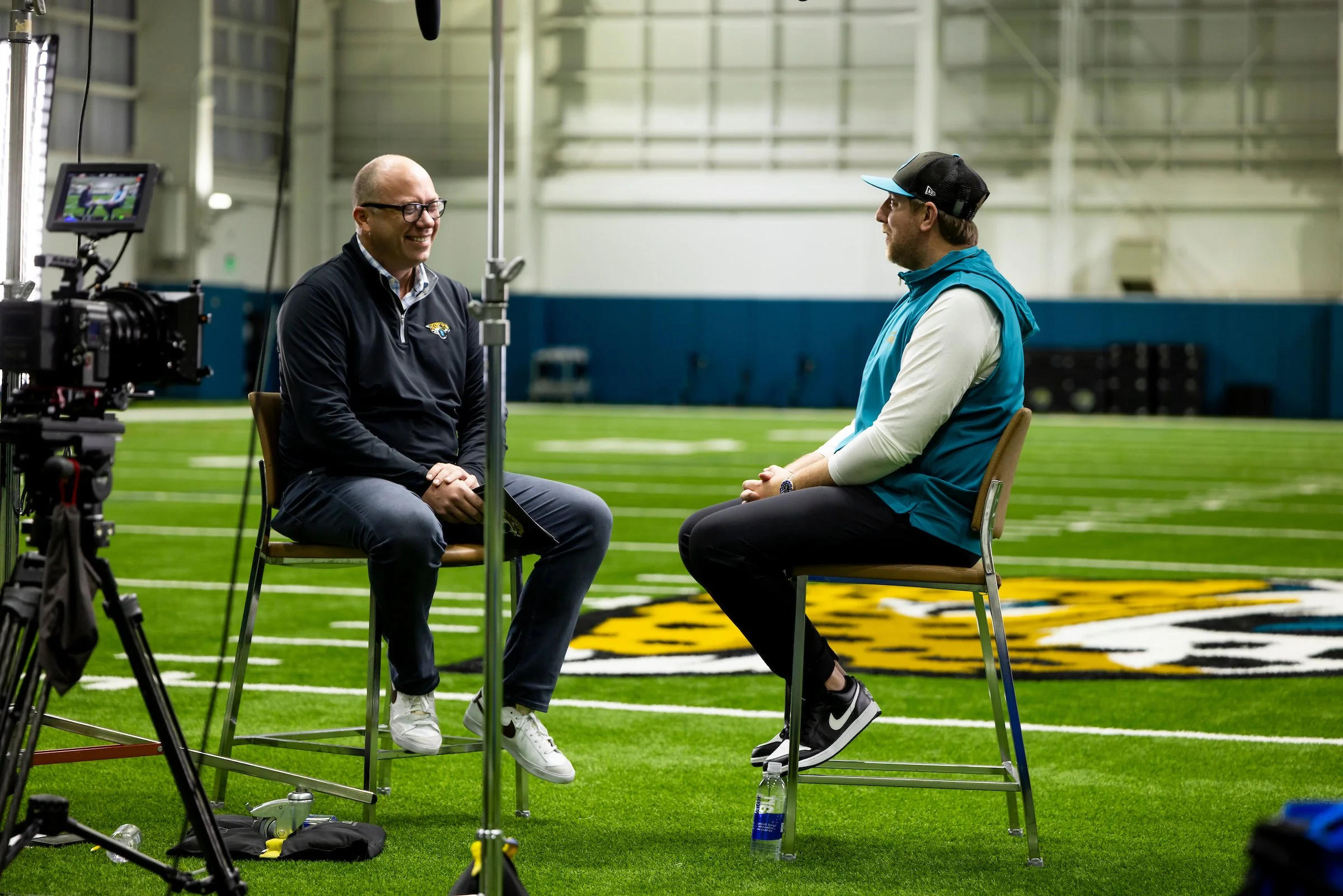 Two men sitting in chairs facing each other on an indoor football field, recording an interview with camera equipment nearby.