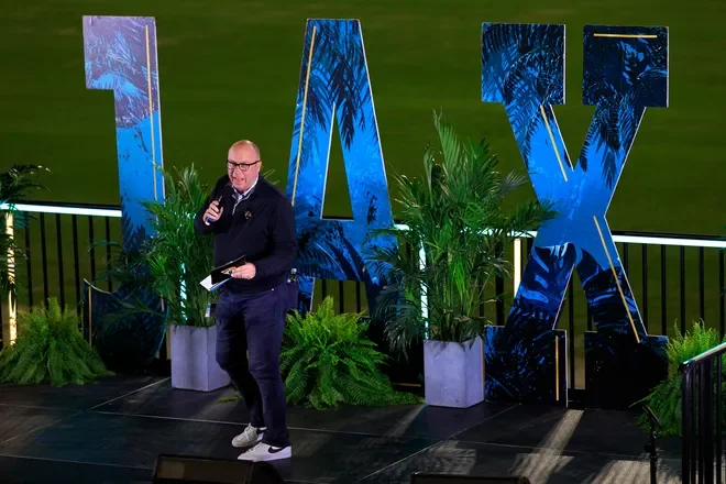 A man speaking on stage at TX event, holding a microphone and notes, with large blue 'TX' letters and green plants in the background.