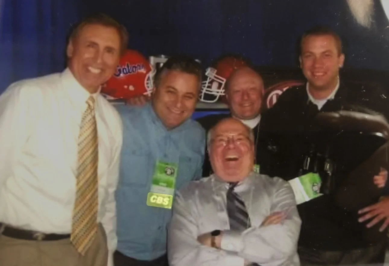 Group of five smiling men at an event, with football helmets in background