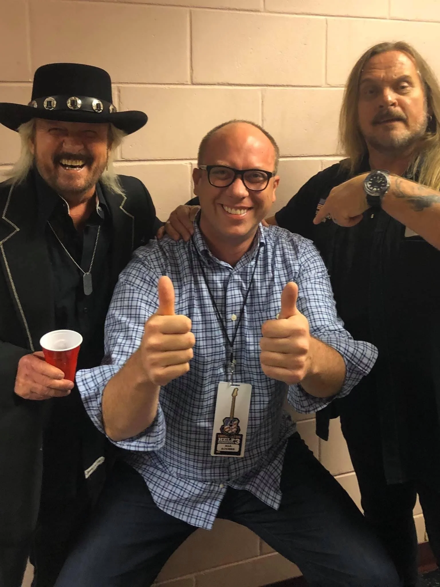 Three men smiling and posing for a photo against a brick wall, with one giving a thumbs-up and another pointing at his wristwatch. The man in the middle wears glasses and a checkered shirt, while the man on the left has a large hat and holds a red cu