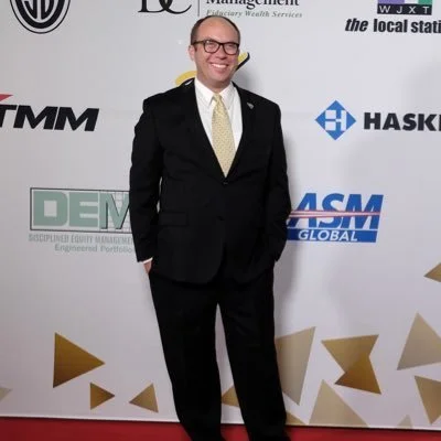 A man wearing a black suit, light-colored shirt, and yellow tie standing on a red carpet in front of a step and repeat banner with various logos.