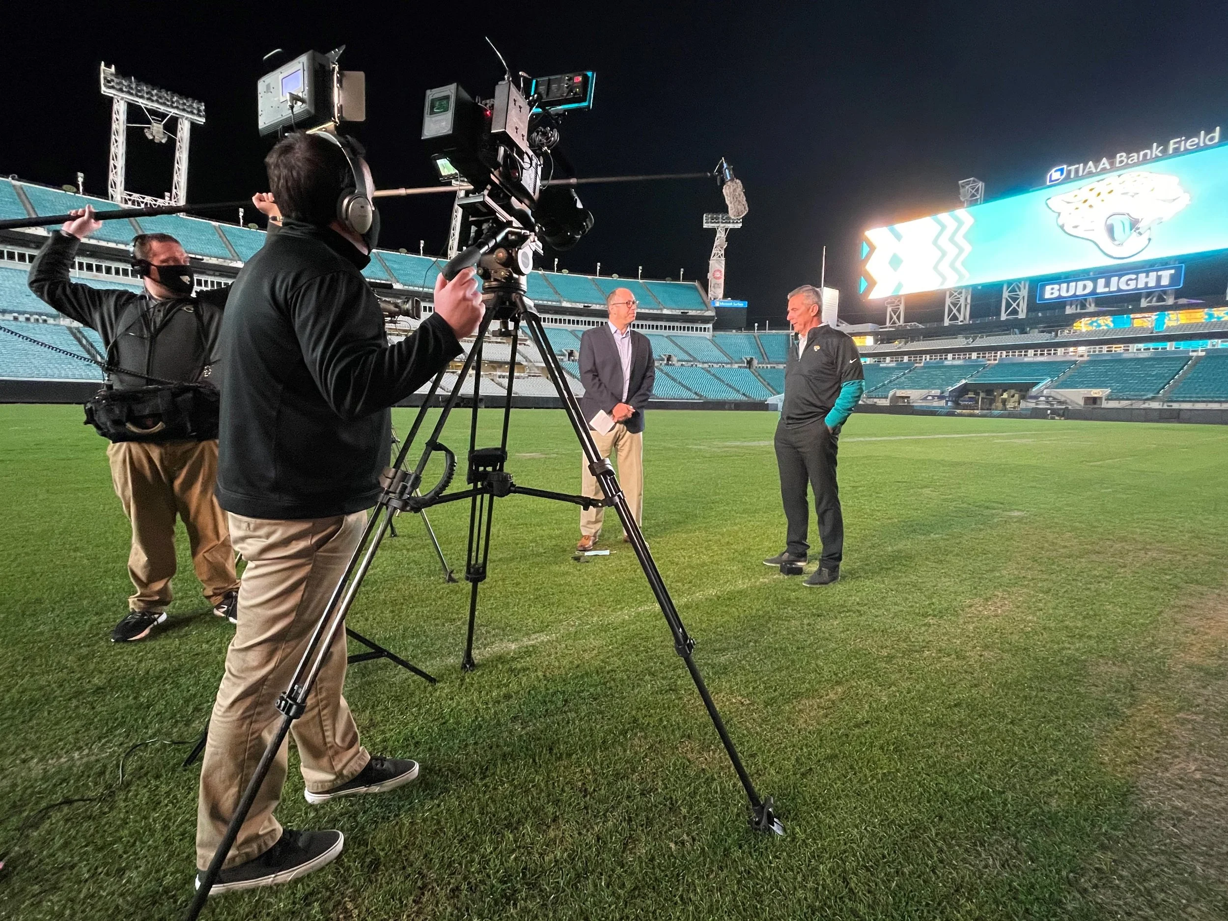 A television crew is recording an interview on a football field at night. The scene includes a cameraman operating a camera, two men standing and speaking, and a large electronic scoreboard displaying a jaguar logo and advertisements.