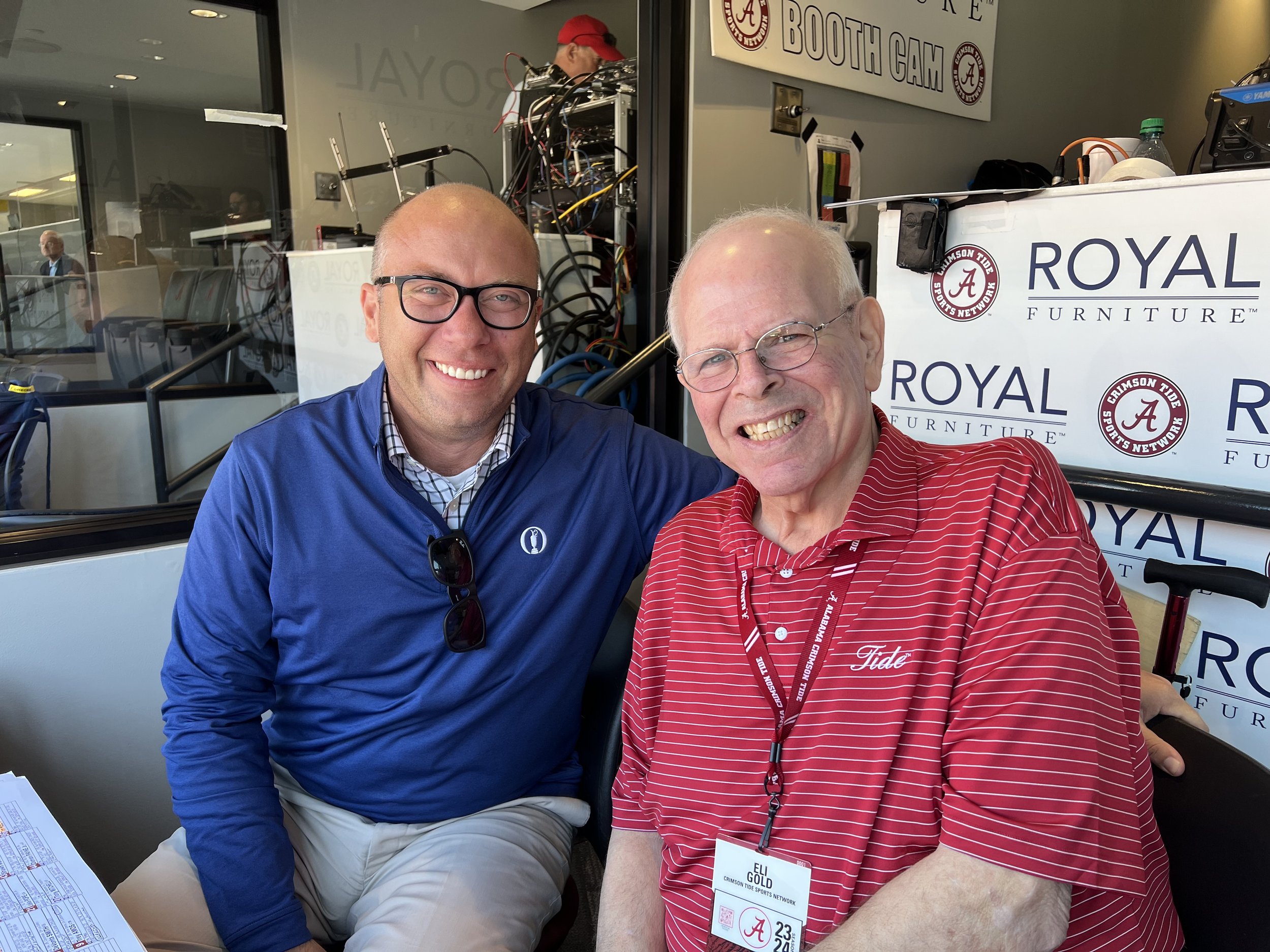 Two smiling men sitting close together at a table, one in a blue jacket and glasses, the other in a red striped shirt and glasses, with event lanyards around their necks, in front of a backdrop with 'Royal Furniture' and 'Crimson Tide Sports Network'