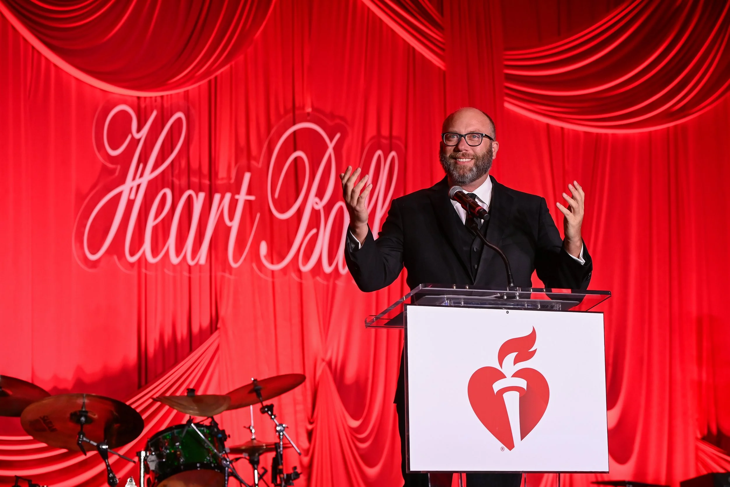 A man with glasses, a beard, wearing a black suit and tie, stands behind a podium giving a speech at an event with red curtains in the background. The podium has a heart and flame logo. The backdrop has the words 'Heart Ball' written in cursive.
