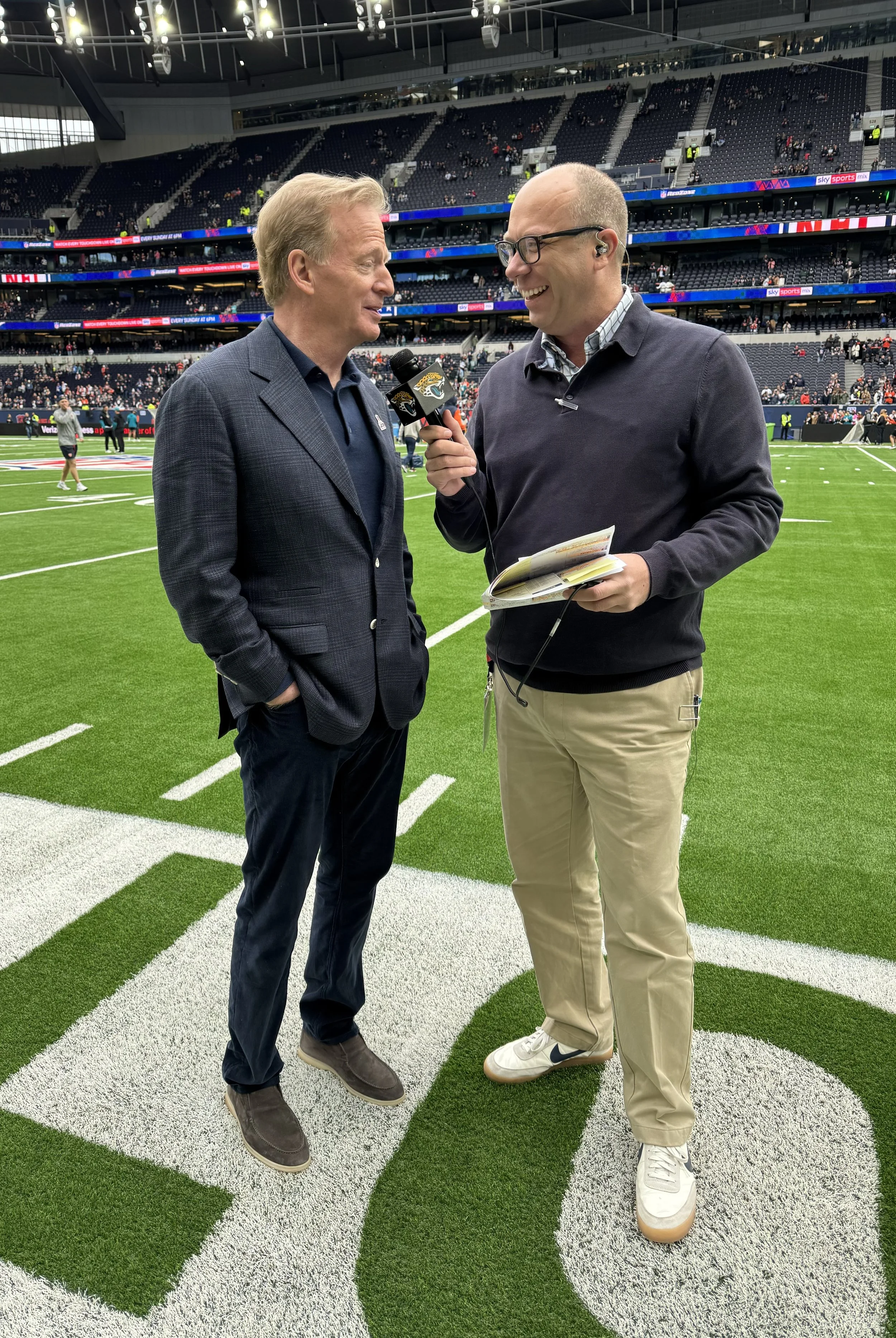 Two men standing on a football field engaging in an interview, one holding a microphone with a Jaguars logo, the other with a notepad, in a stadium with empty seats and people in the background.