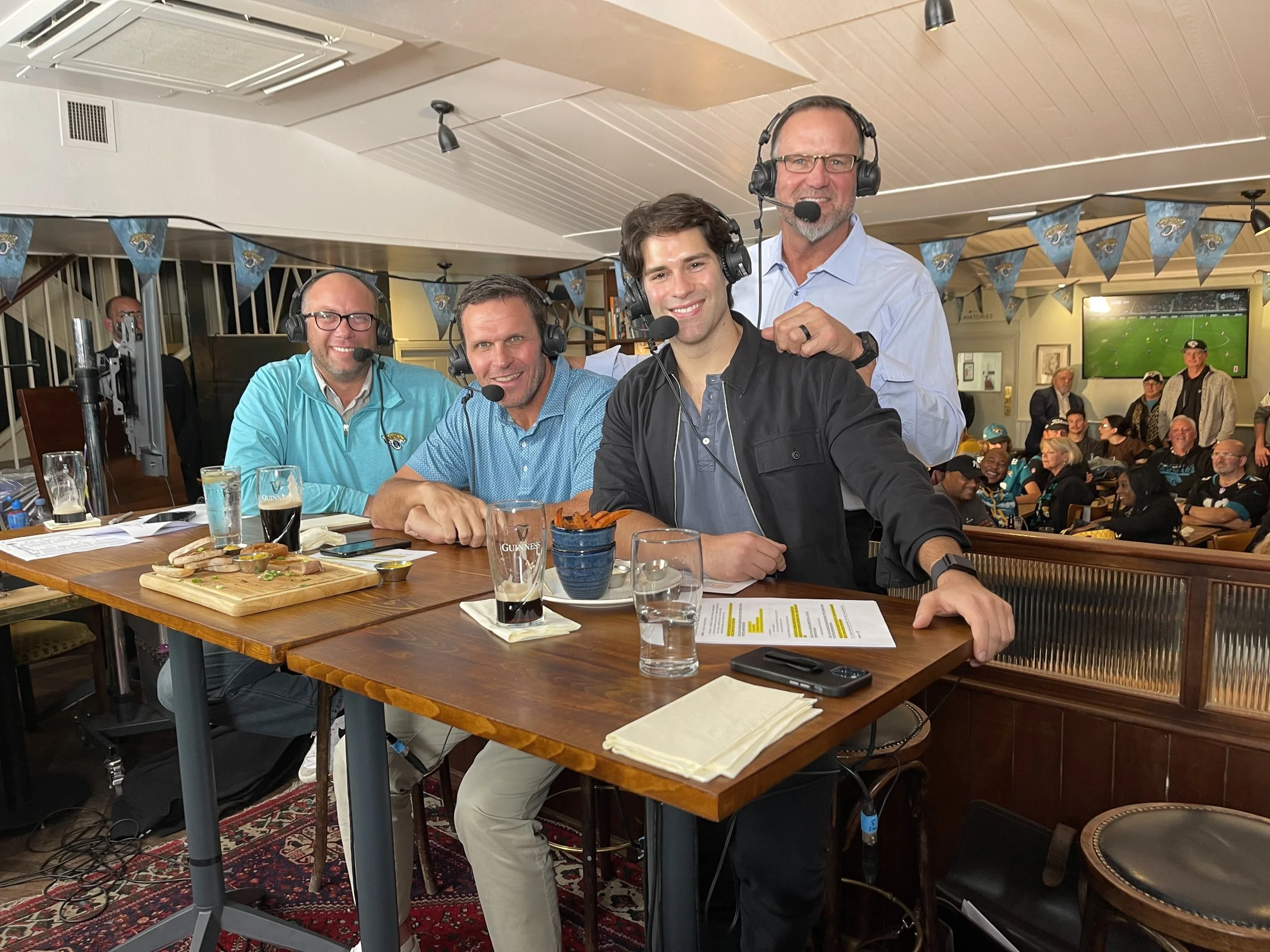 Group of four men wearing headsets and sitting at a table with drinks and snacks, with an audience watching and a TV in the background, at a sports bar or restaurant.