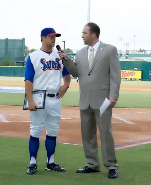 A baseball player in a Suns uniform being interviewed by a man in a suit on the baseball field.