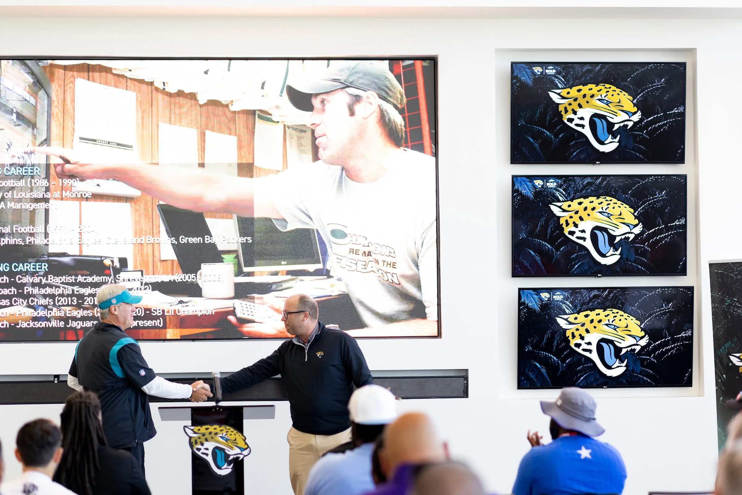 Two men shake hands at a podium featuring the Jacksonville Jaguars logo during a press event, with an audience in front. Large monitor displays a man in a cap pointing, with text about his football career. Three smaller screens show the Jacksonville 