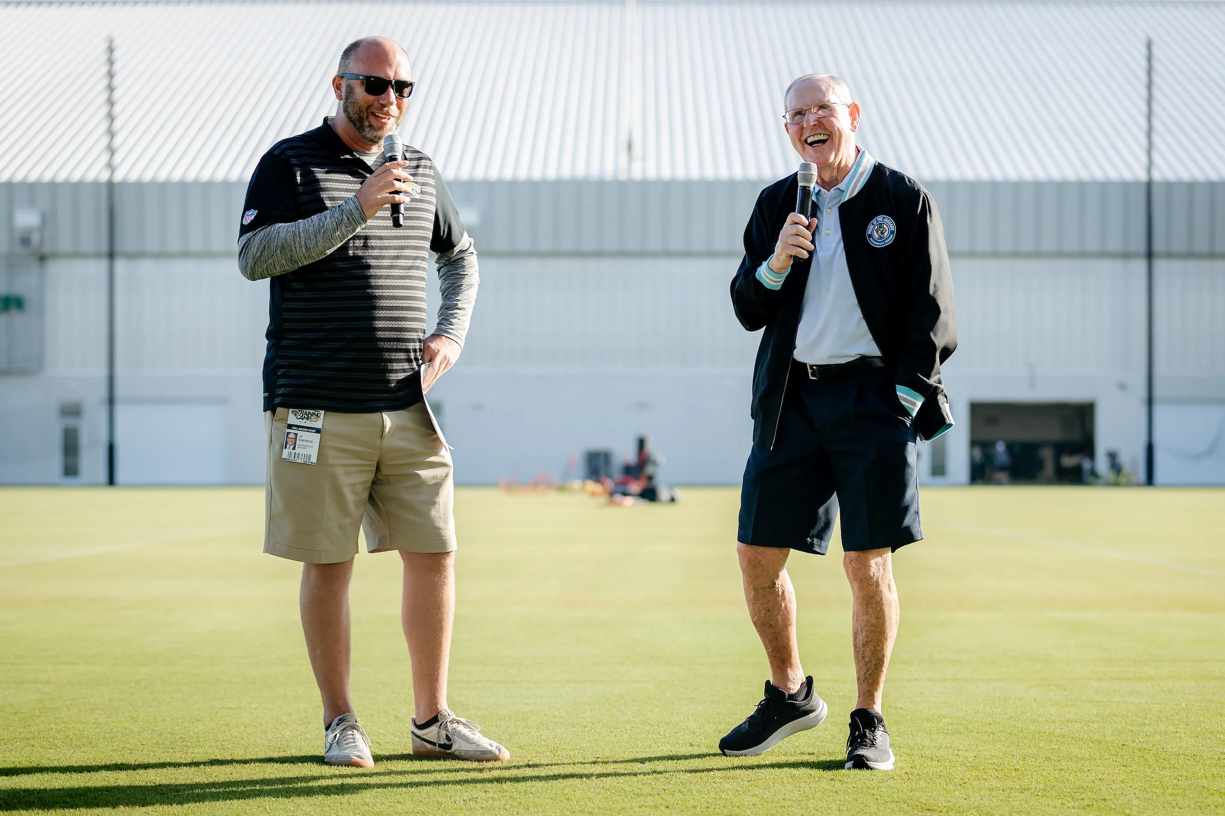 Two men standing on a grassy field outdoors, each holding a microphone and speaking. One man is wearing sunglasses, a striped long-sleeve shirt, shorts, and sneakers. The other man is wearing glasses, a jacket, shorts, and sneakers, and appears to be
