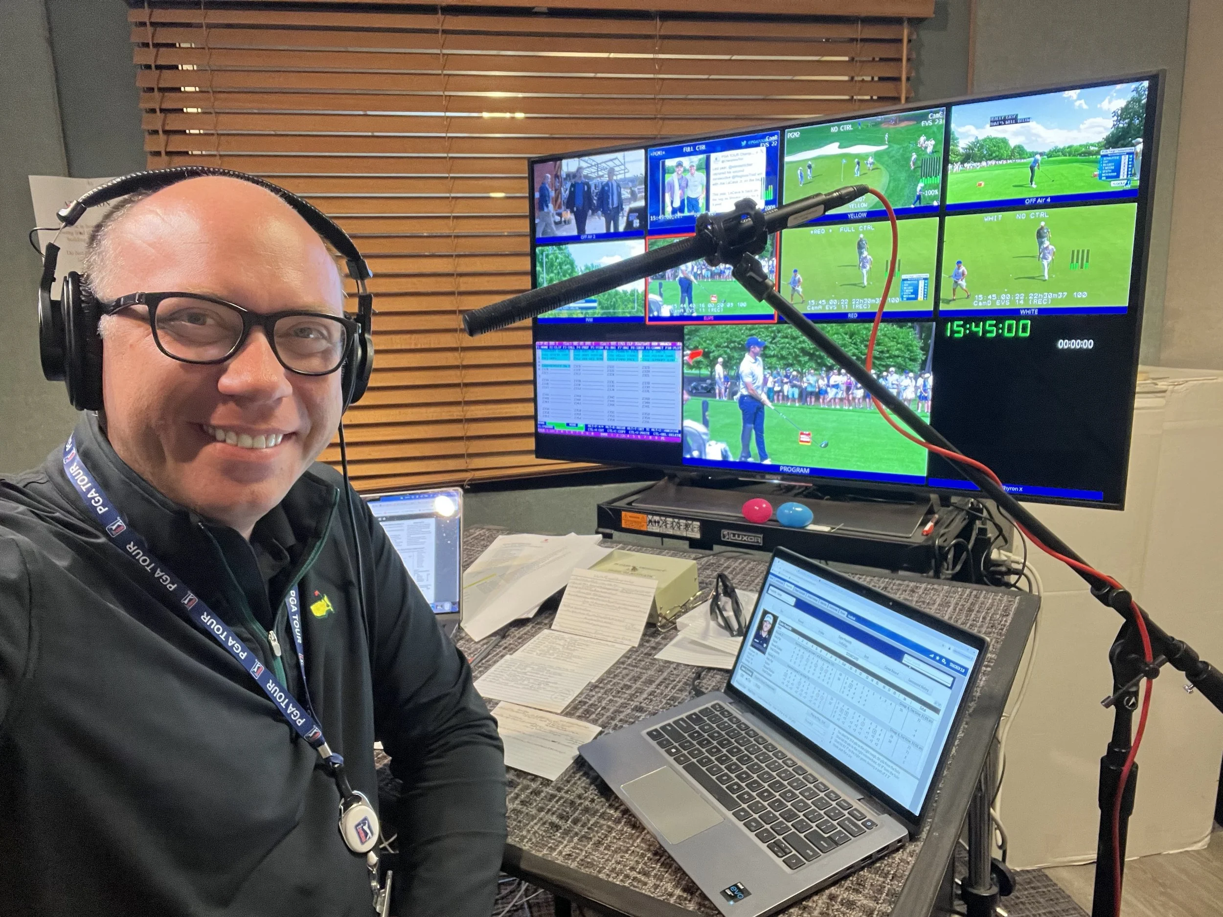 A smiling man wearing glasses and headphones sitting at a desk with multiple laptops, papers, and documents, in front of a large monitor displaying multiple sports and camera feeds.