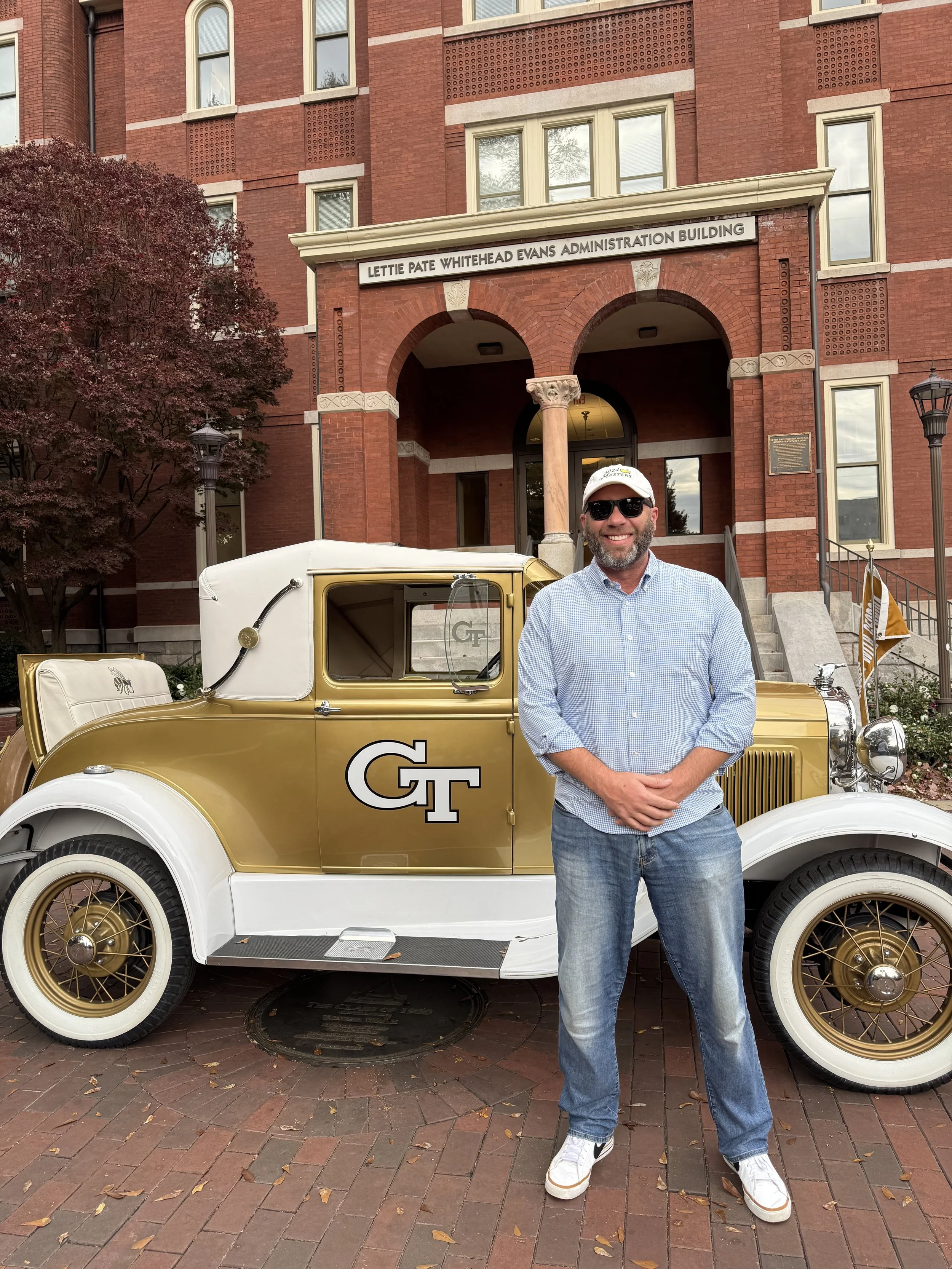 A man standing in front of a vintage gold and white car with a Georgia Tech logo on the door, in front of a brick building labeled 'Lettie Pate Whitehead Evans Administration Building'. The man is wearing sunglasses, a white cap, a light blue checked