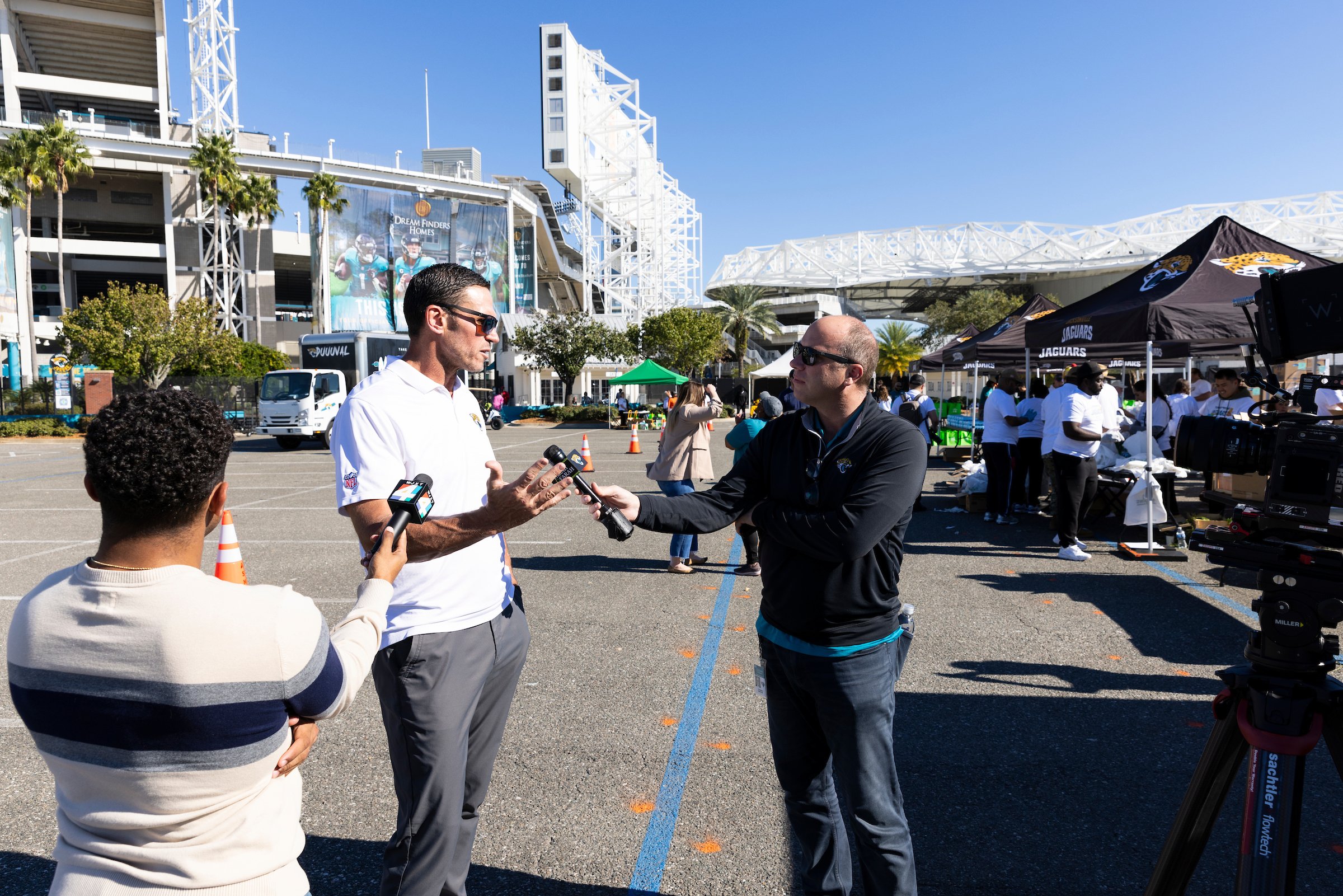A man being interviewed by a reporter at an outdoor event near a stadium, with several tents and people in the background.