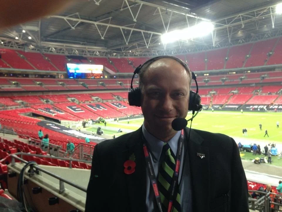 A man wearing a suit and tie with a poppy pin, headset, and lanyard, standing in a sports stadium with red seats and a field in the background.
