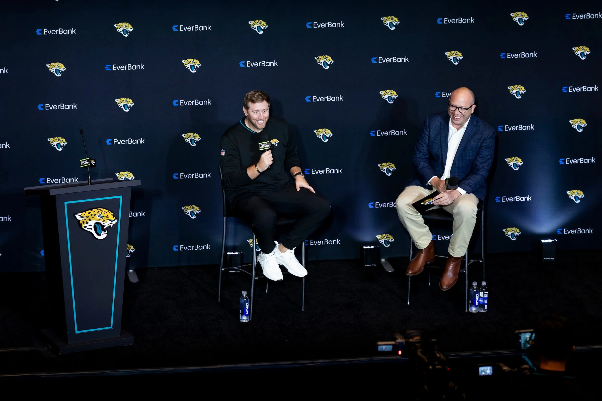 Two men sitting on stools and laughing during a panel at an EverBank event, with a black backdrop featuring the Jacksonville Jaguars logo and EverBank branding.