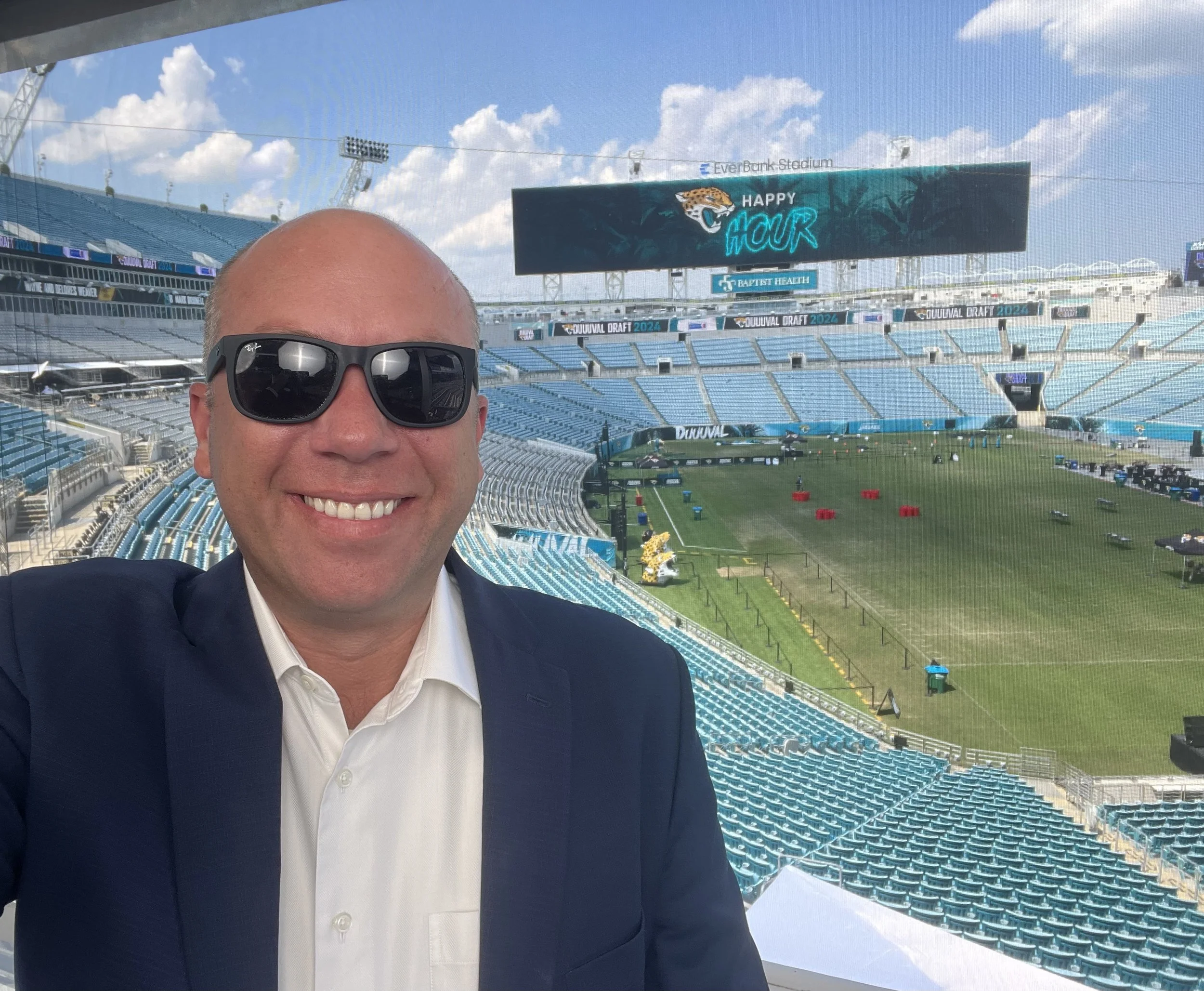A smiling man in sunglasses and a suit taking a selfie at a football stadium, with an empty field and seating in the background under a partly cloudy sky.