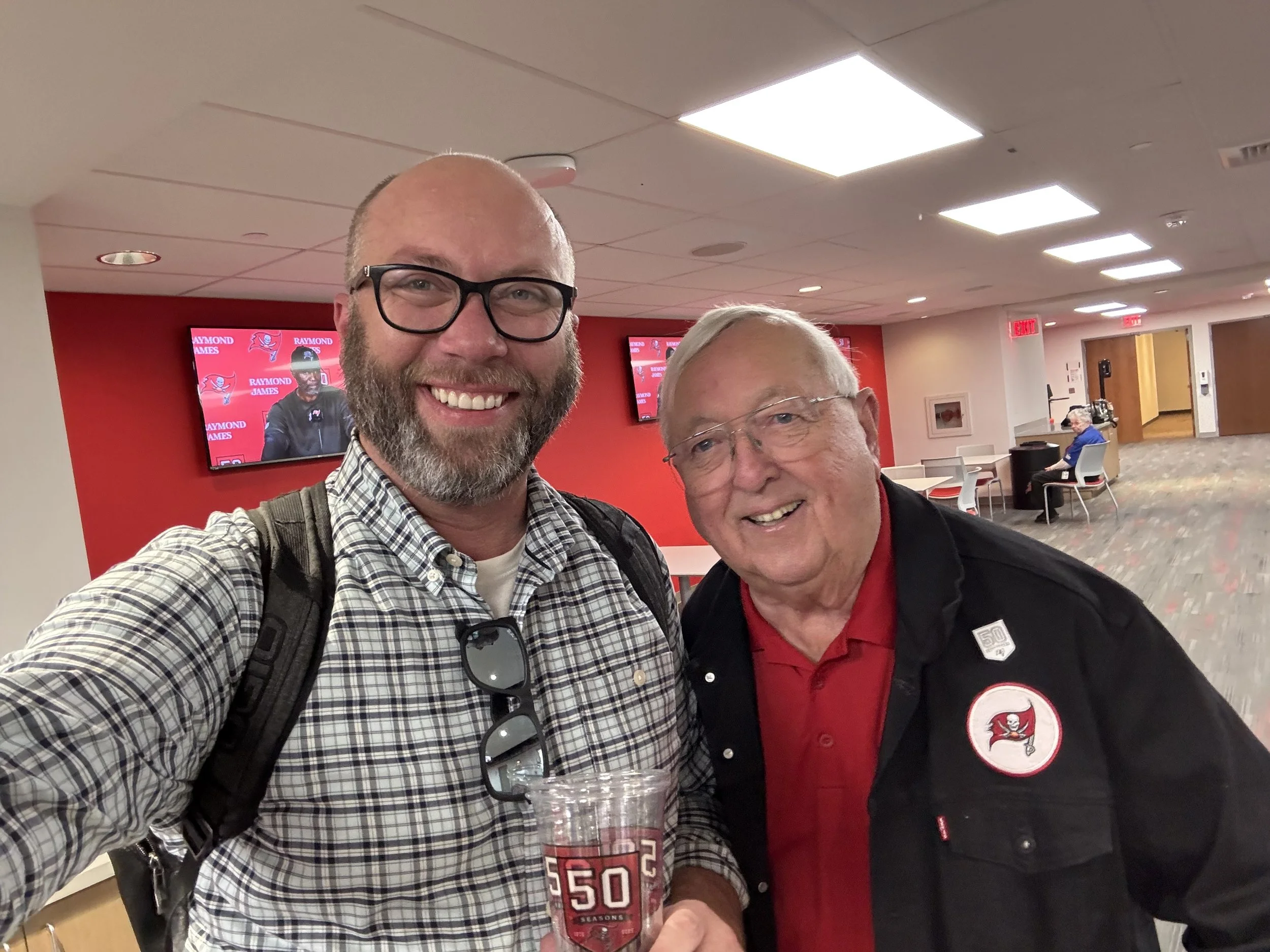 Two smiling men taking a selfie in an indoor lounge area with chairs and televisions. One man has a beard, glasses, and a checkered shirt, holding a clear cup with the number 50 on it. The other man has glasses, gray hair, and is wearing a black jack