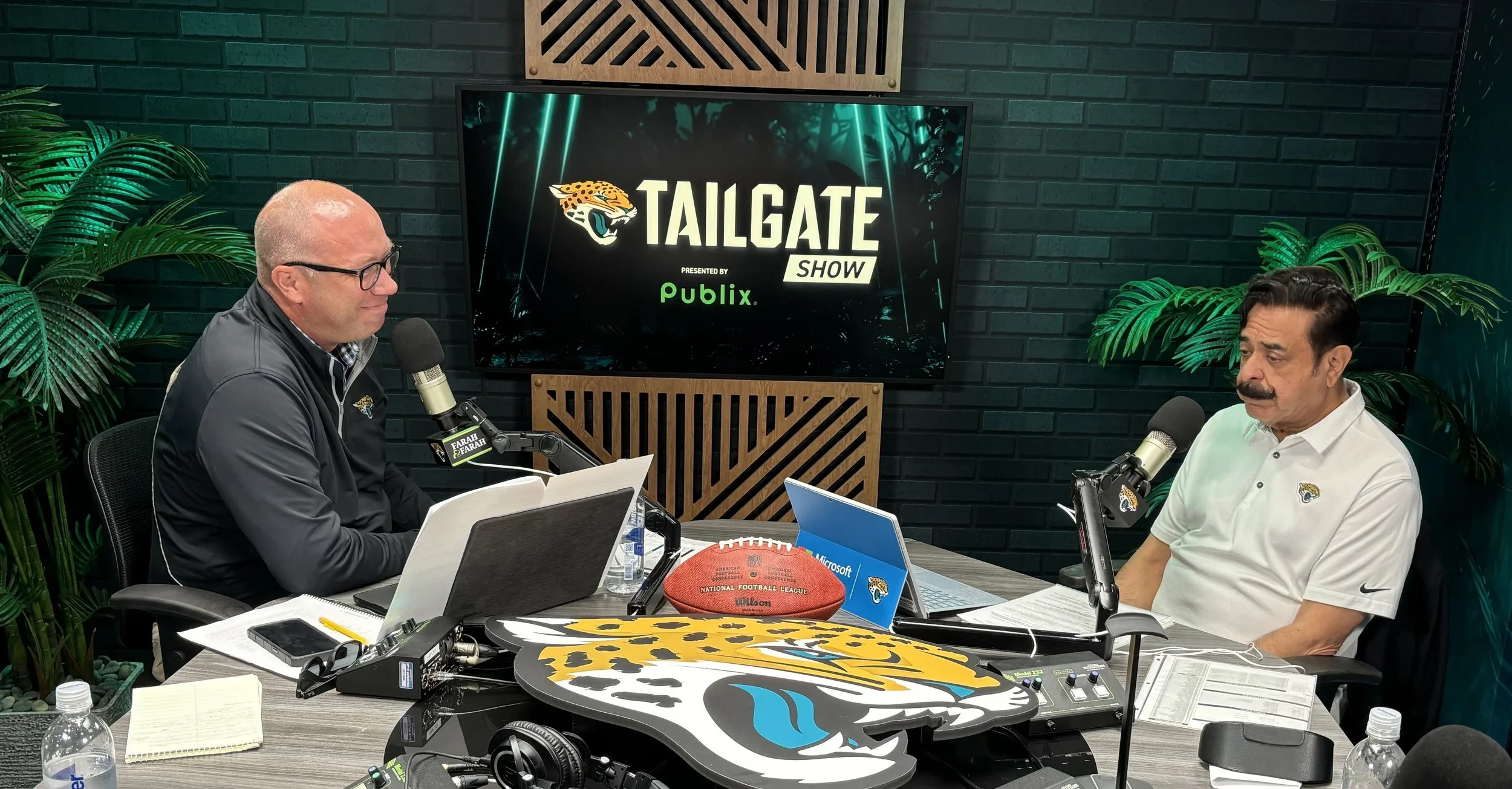Two men sitting at a table with microphones, laptops, and notes, engaged in a discussion in a radio studio with a Jaguars theme, including a large Jaguars logo on the table and a television screen behind them that reads 'Tailgate Show' presented by P