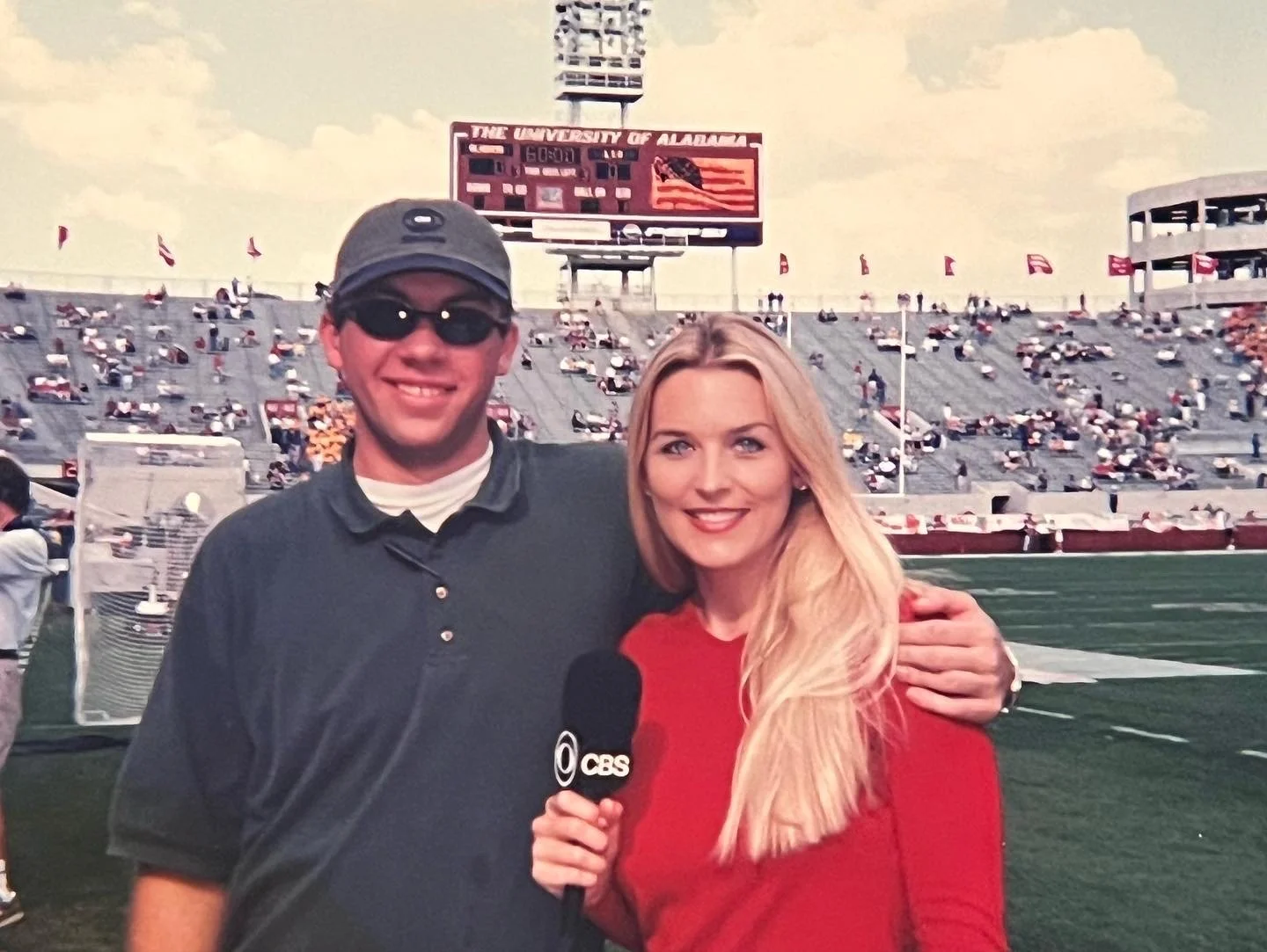 A man and a woman at a football stadium, with the man holding a CBS microphone. The man wears sunglasses, a cap, and a dark polo shirt. The woman has long blonde hair, wears a red top, and is smiling. The stadium seats and a scoreboard are visible in