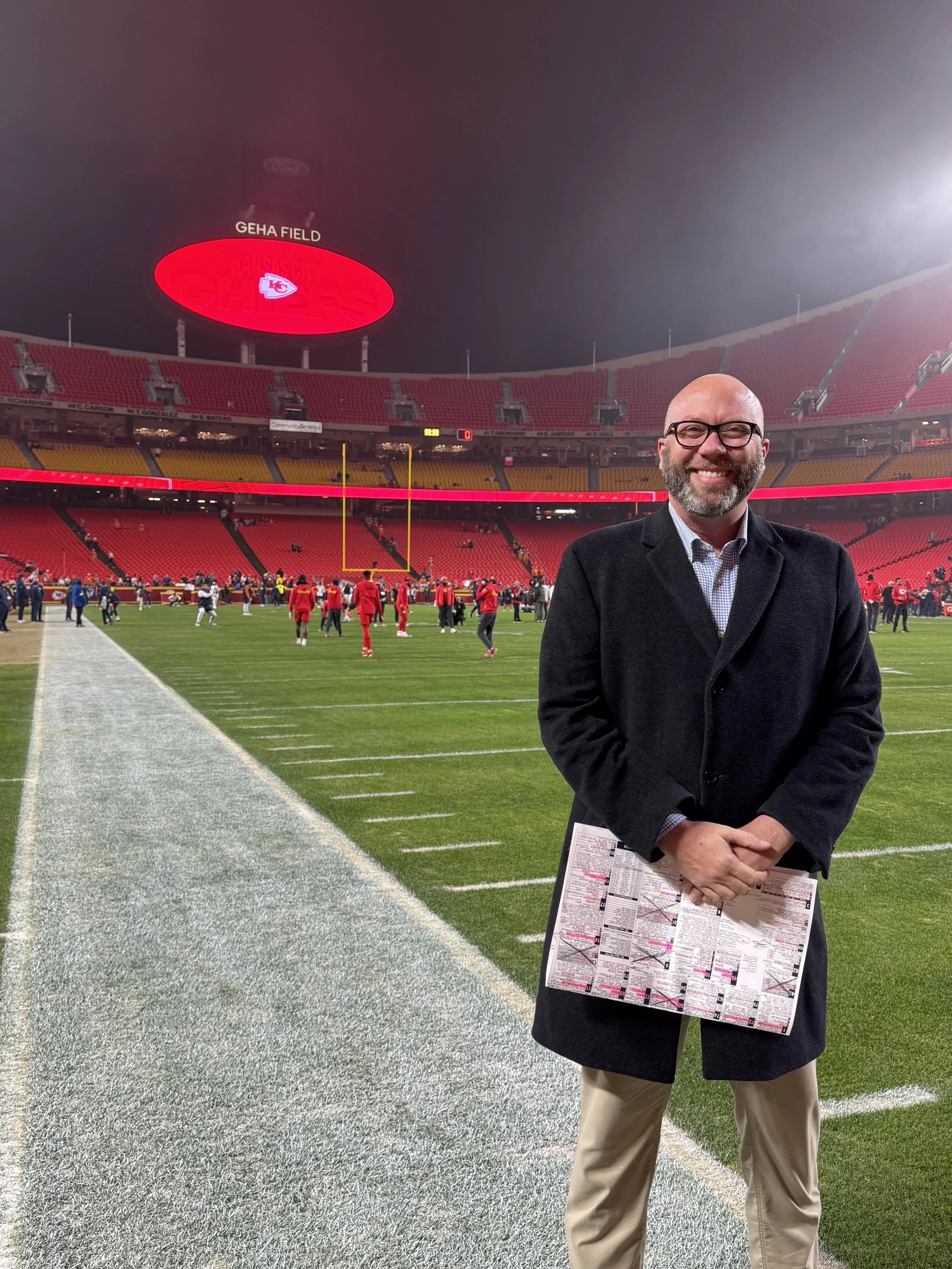 A smiling man in glasses and a dark coat standing on a football field at Arrowhead Stadium at GEHA Field, with players warming up in the background and the stadium mostly empty.
