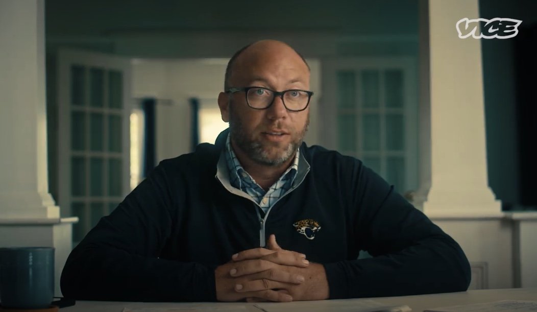 A man wearing glasses and a Jacksonville Jaguars jacket sitting at a table inside a house, with a blue mug nearby.