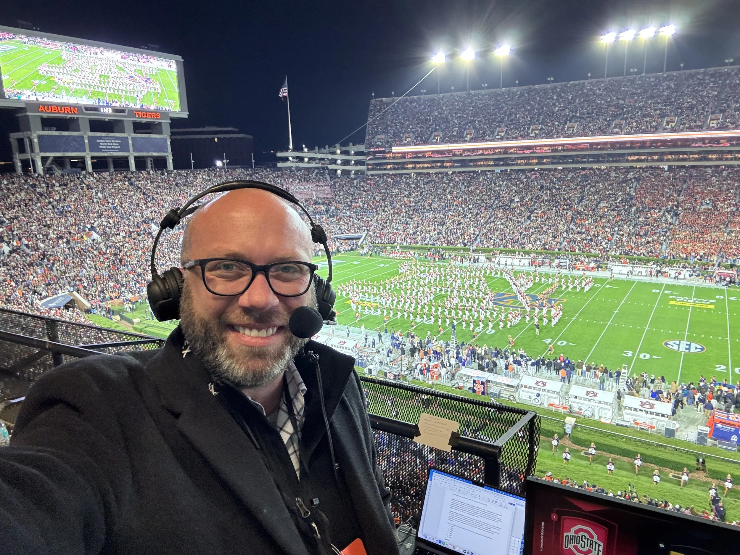 A man with glasses and a headset smiling at the camera, taking a selfie at a football stadium filled with spectators during a game or event at night. The field is visible in the background with players, and a large screen displays a marching band or 