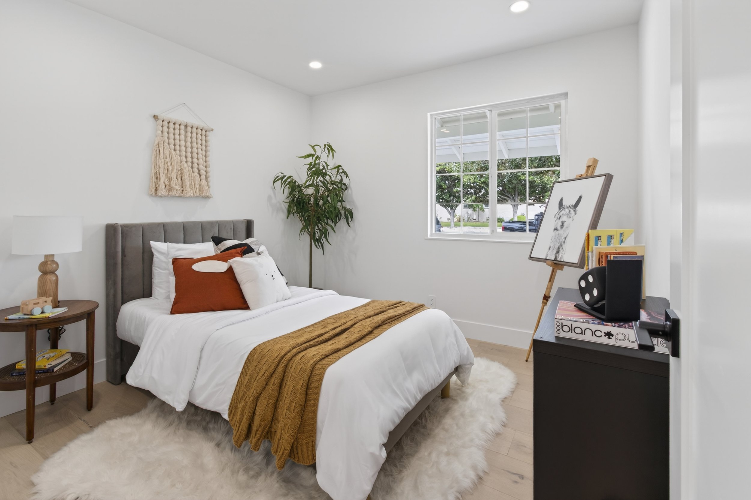 Walnut Creek A bedroom with a bed, pillows, a white fluffy rug, a bedside table with books, a lamp, a potted plant near the window, and an easel with a llama drawing, with decor on the wall and a window showing greenery outside.