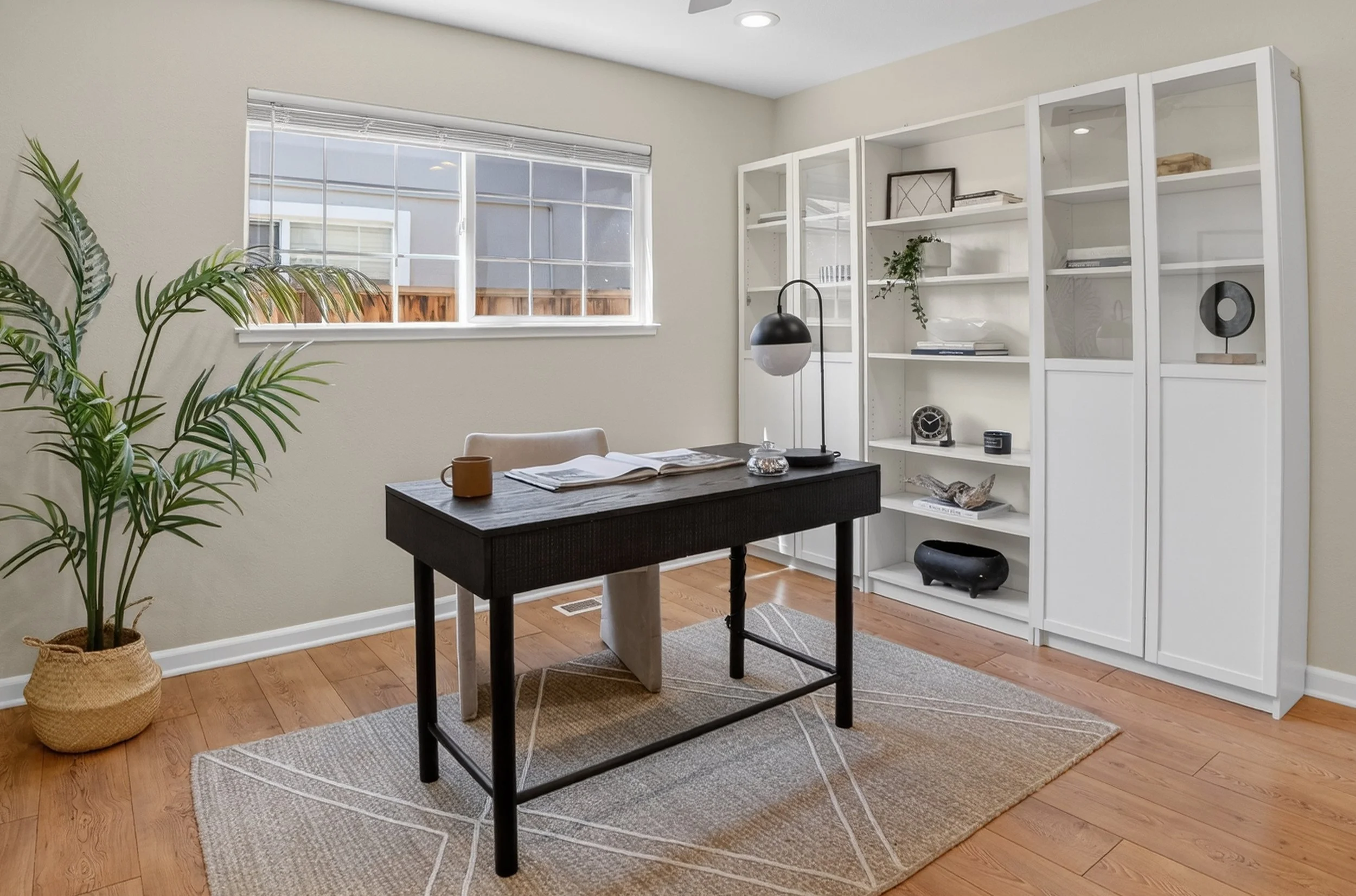 Home office with a black desk, open book, coffee mug, black and white table lamp, white bookshelf with decorations, large window, wooden floor, and a potted plant.