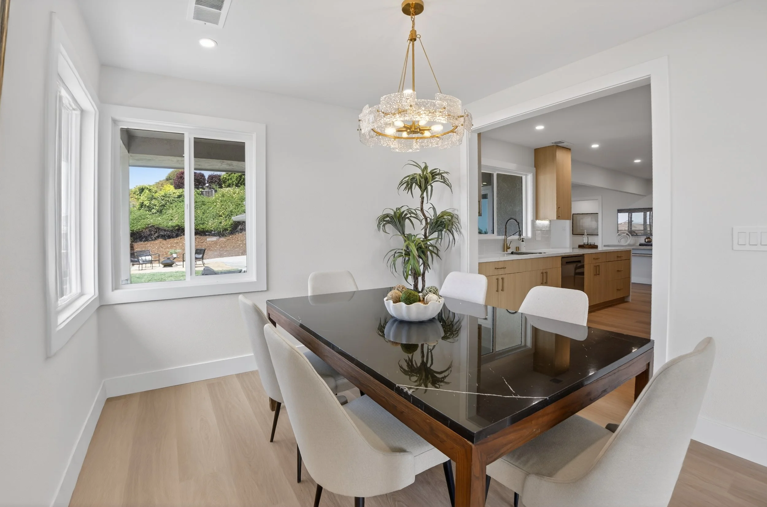 Dining room with a rectangular black marble table, six white upholstered chairs, a chandelier, and a large window with a view of a backyard with chairs and greenery. Open kitchen with wooden cabinets and a sink in the background.