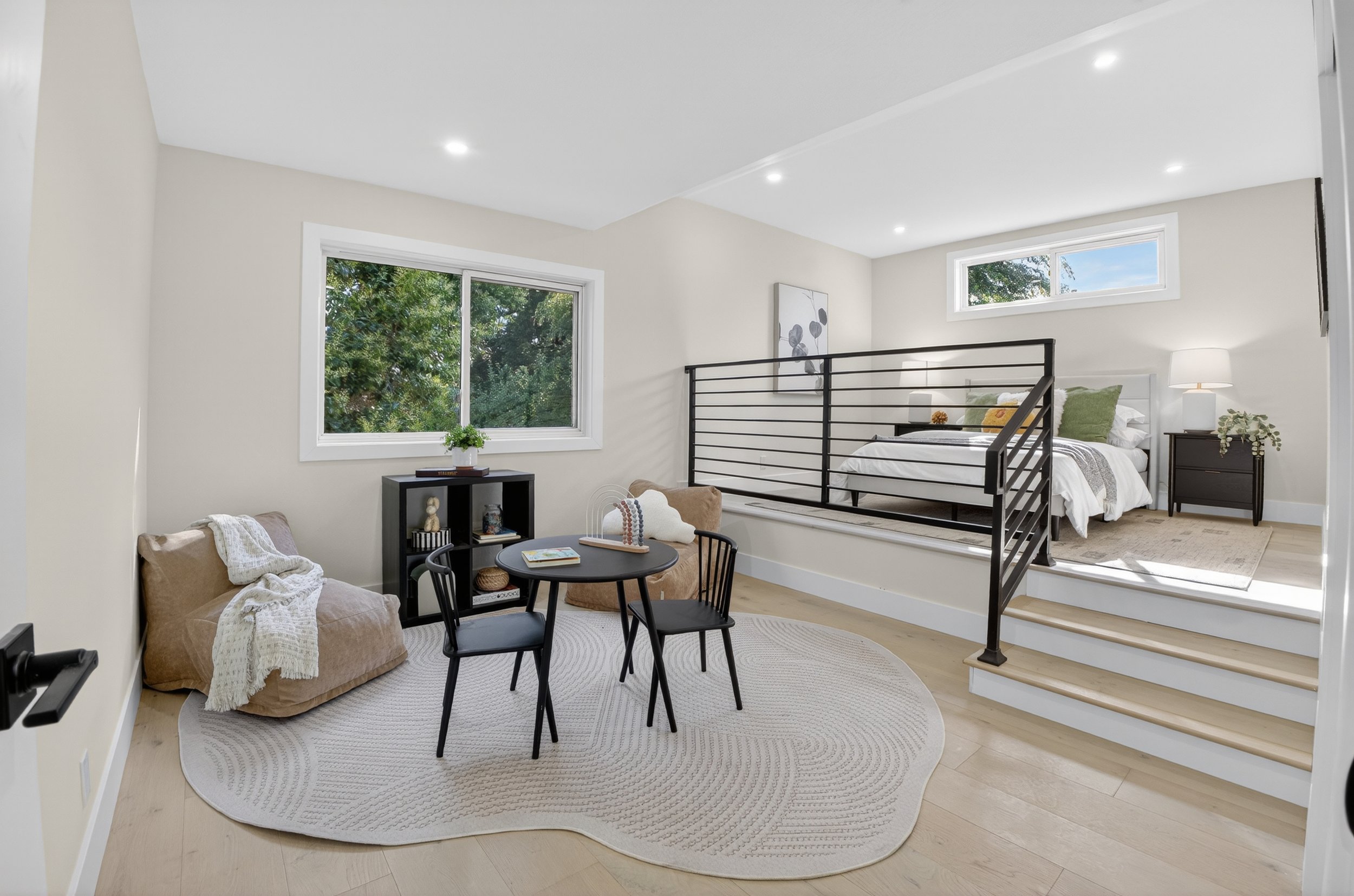 A modern bedroom with a seating area, black metal railing, and a bed with white linens and green pillows, illuminated by natural light from windows.