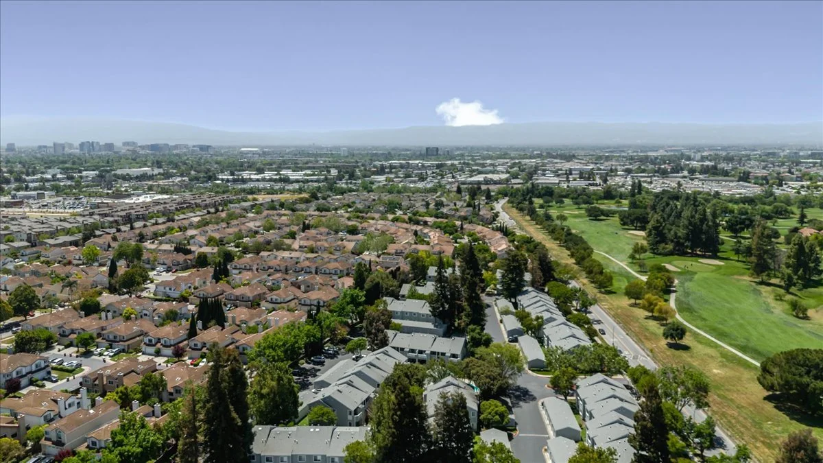Aerial view of a suburban neighborhood next to a large green park with walking paths and trees, with a city skyline and mountains in the distance.