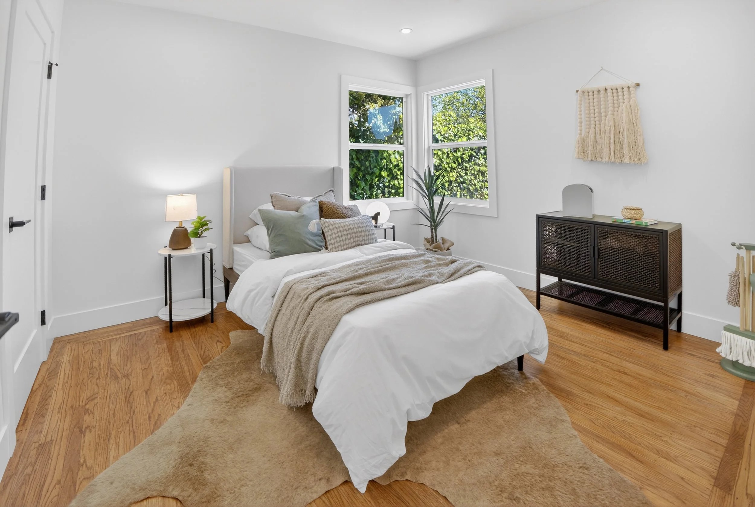 A cozy bedroom with a white bed, throw blanket, multiple pillows, hardwood floors, a side table with a lamp and plant, large windows letting in natural light, a black cabinet, and wall decor.