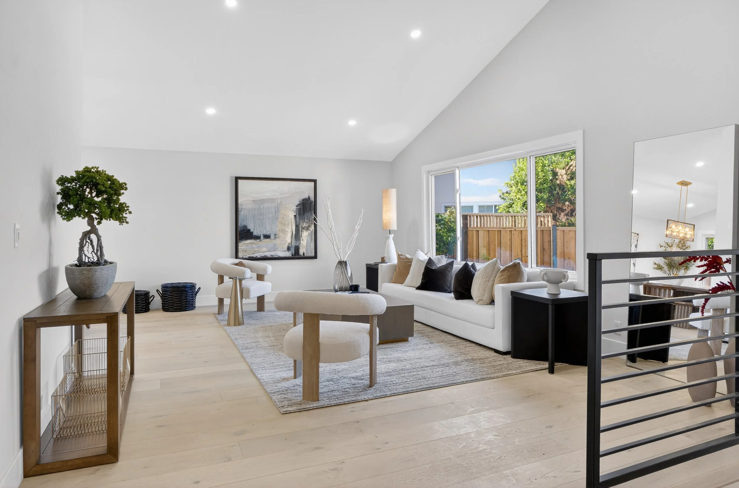 Bright living room with white walls, wood flooring, a large window, a white sofa with beige, black, and white pillows, a small black side table, a textured area rug, a wooden console with a bonsai tree, a framed abstract painting, decorative vases, a