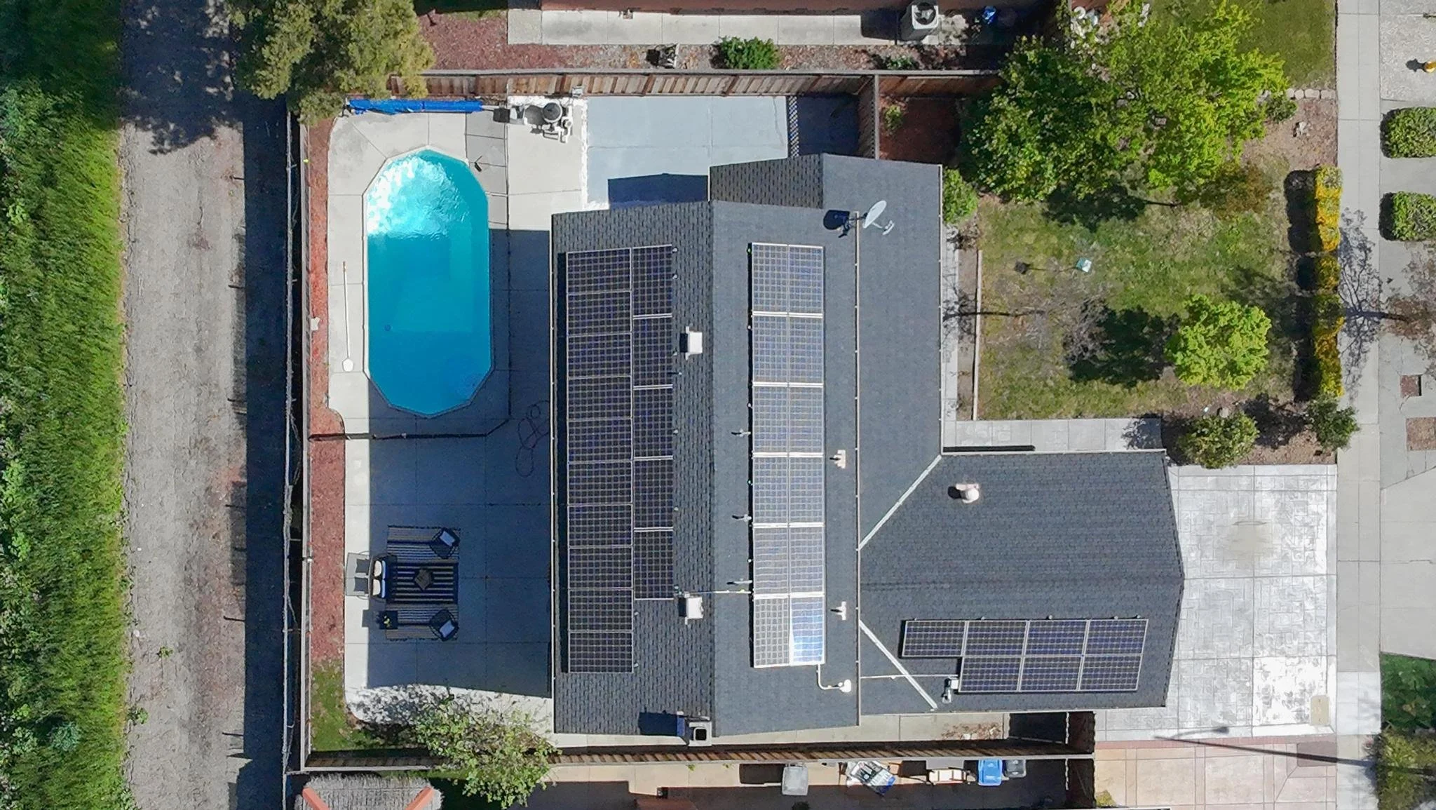 A house roof with solar panels and a small swimming pool in the backyard, surrounded by trees and a fenced yard.
