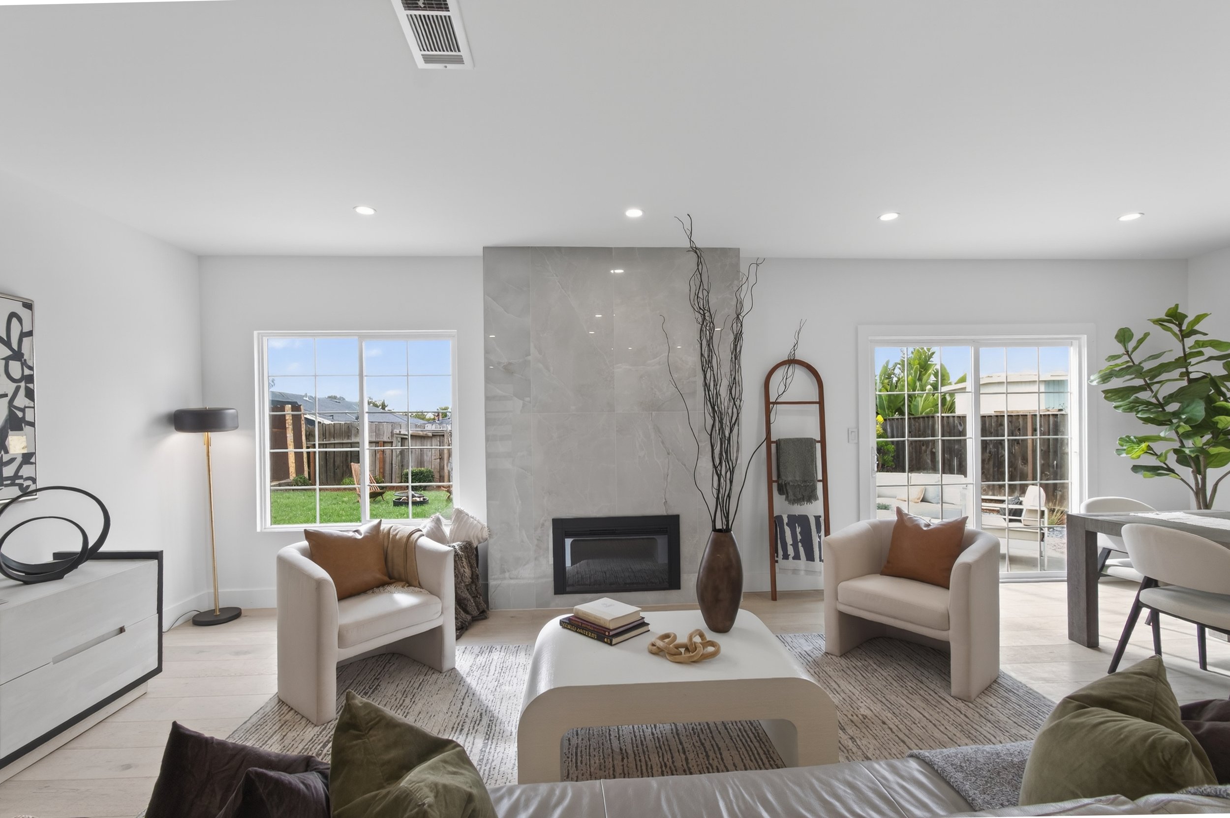 Living room with white walls, a fireplace, and sliding glass doors leading outside. Furniture includes two white armchairs with brown pillows, a white coffee table with books and decorative items, and a sofa with pillows. There is a tall vase with br