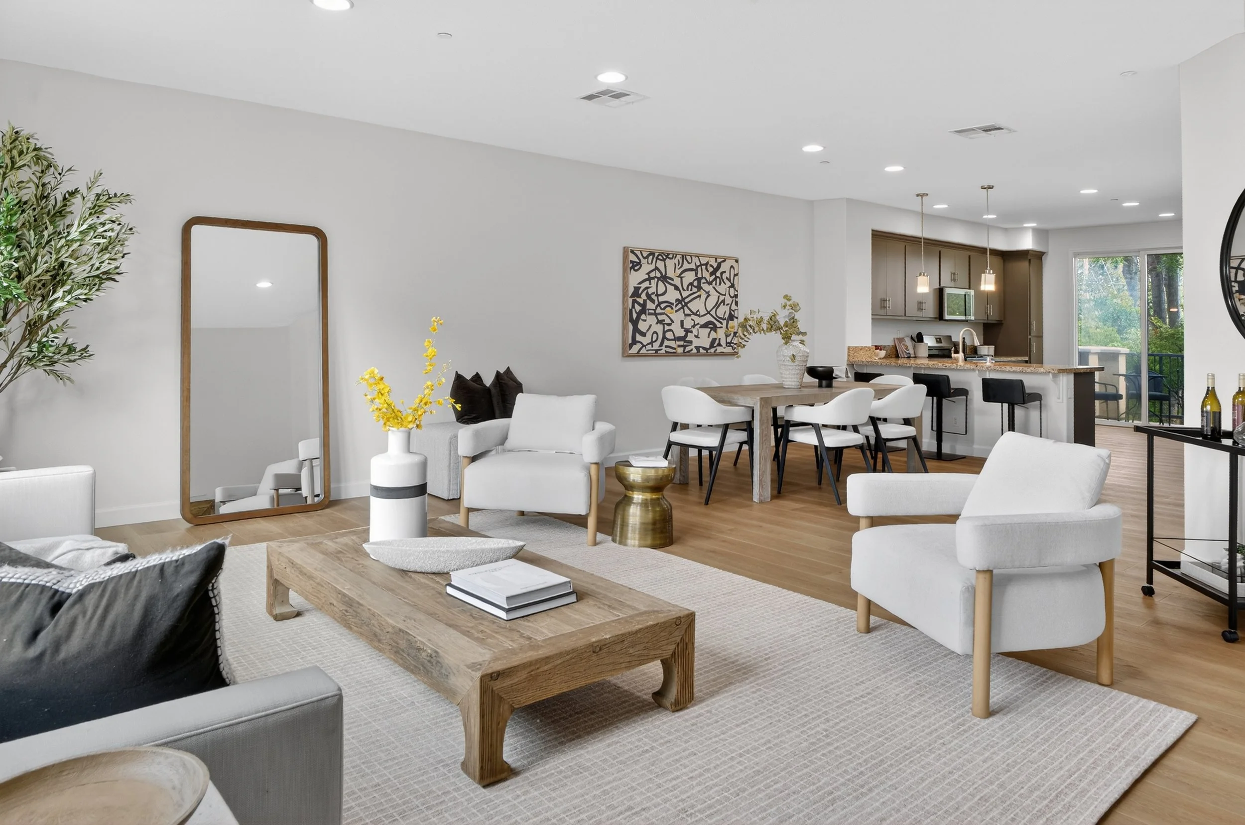 Open-concept living room with white walls, wooden flooring, and modern furniture including white armchairs, a beige sofa, and a wooden coffee table. Decor includes a large mirror, yellow flowers in a white vase, black cushions, and abstract artwork. 