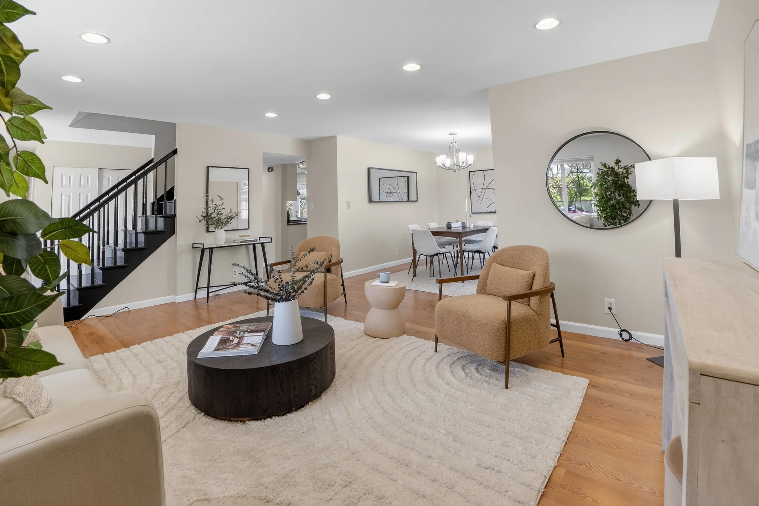 Livermore Living room with beige armchairs, a round wooden coffee table with magazine and vase, area rug, and a sideboard with lamp and mirror, open to dining area with table and chairs.