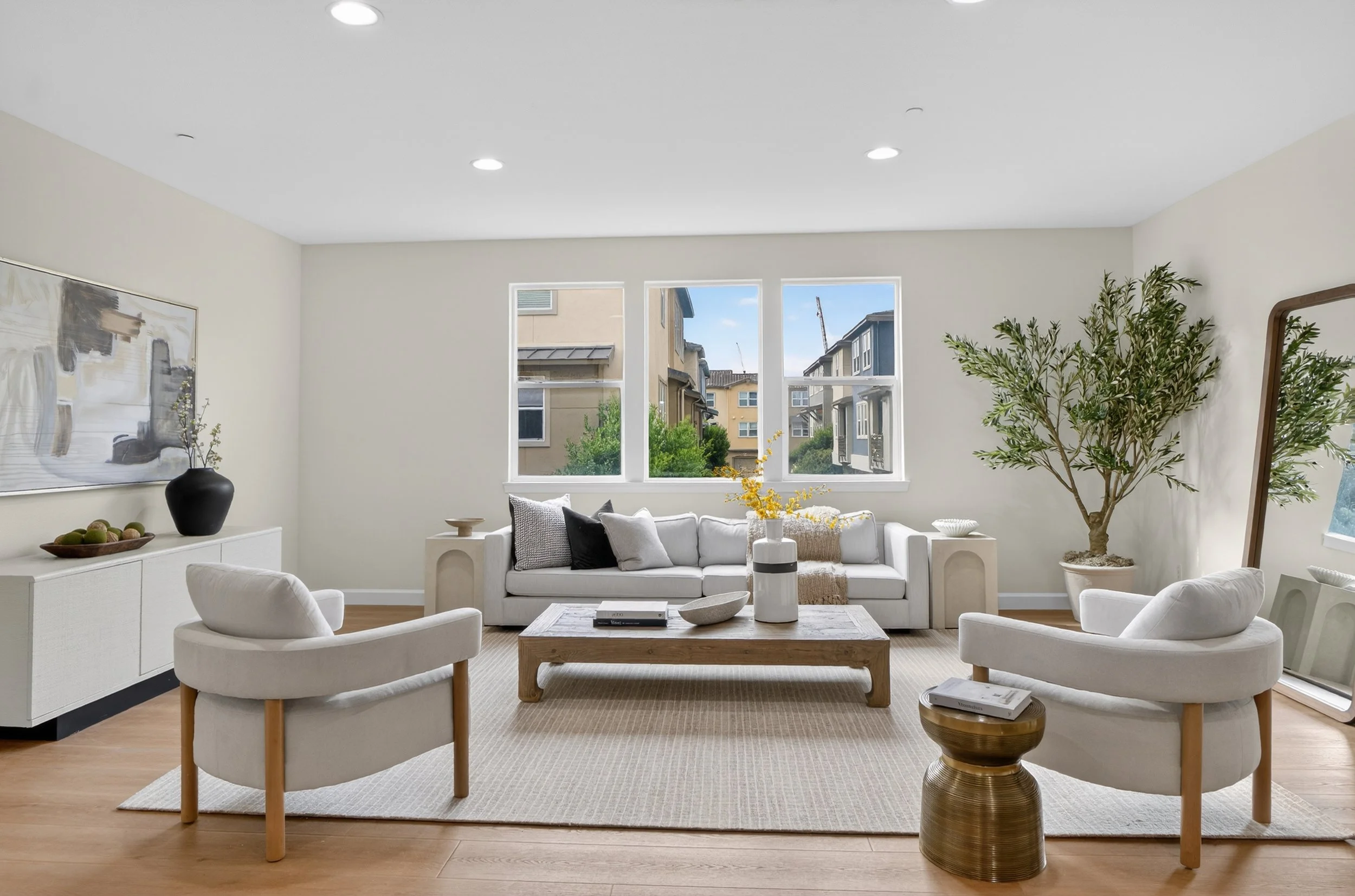 San Jose Modern living room with white sofa, two white armchairs, a wooden coffee table, and large windows showing an apartment complex outside. Decor includes a large black vase, abstract artwork, and potted plant.
