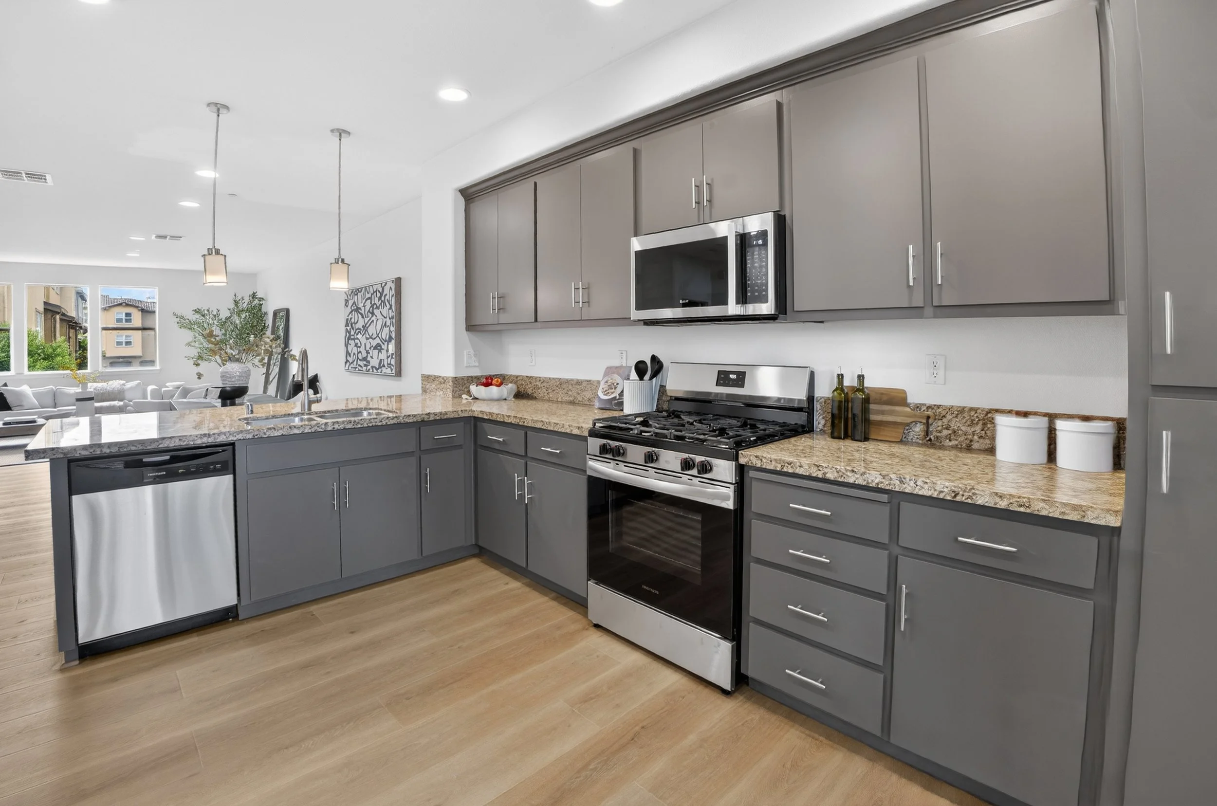 Kitchen with gray cabinets, stainless steel appliances, granite countertops, and wood flooring, opening into a living area with large windows and pendant lighting.