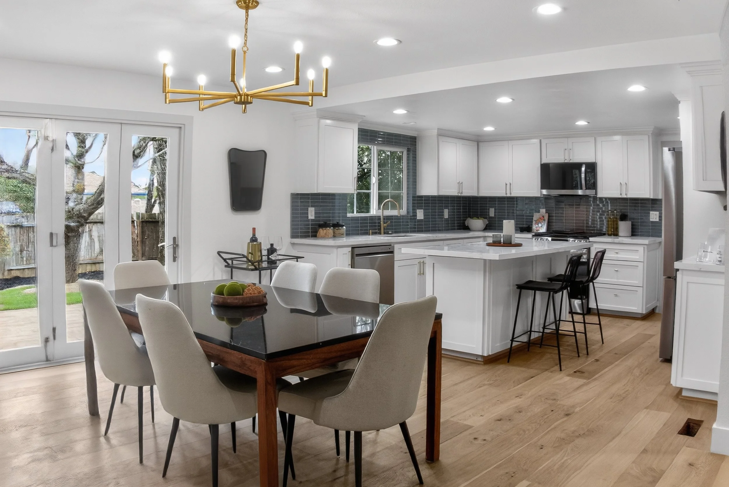 Modern kitchen and dining area with white cabinets, a kitchen island, gray backsplash, stainless steel appliances, a wooden dining table with six beige chairs, and sliding glass doors leading outside.