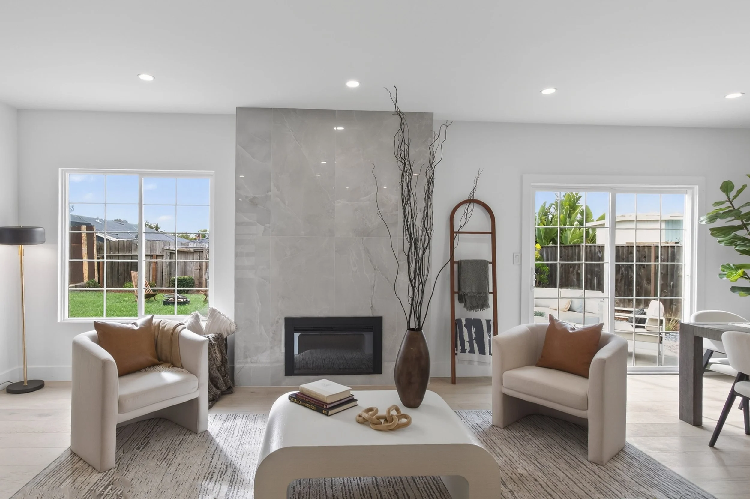 Living room with white walls, a fireplace, and sliding glass doors leading outside. Furniture includes two white armchairs with brown pillows, a white coffee table with books and decorative items, and a sofa with pillows. There is a tall vase with br