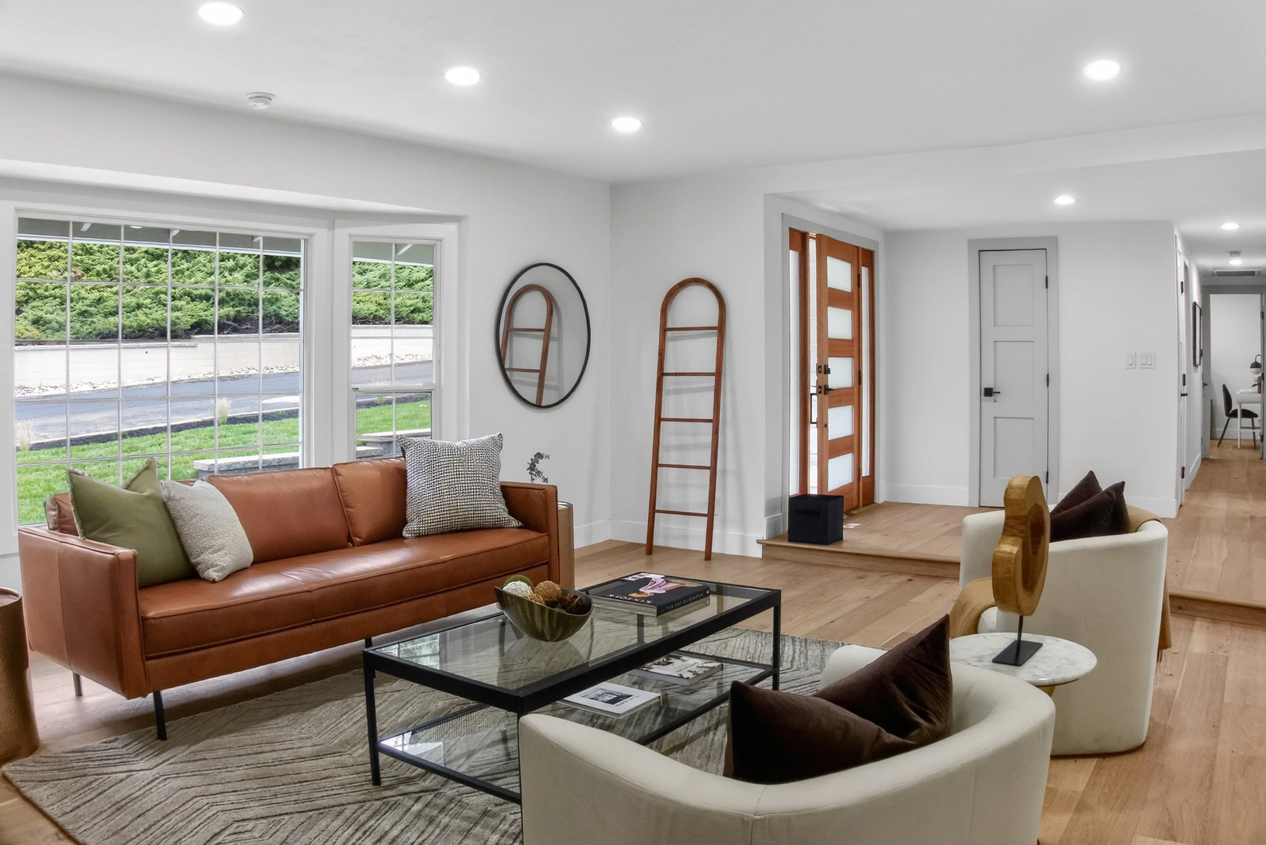 San Ramon Living room with large windows, brown couch with pillows, white armchair with pillows, glass coffee table with books, wooden ladder decor, wooden front door, and view of green landscape outside.