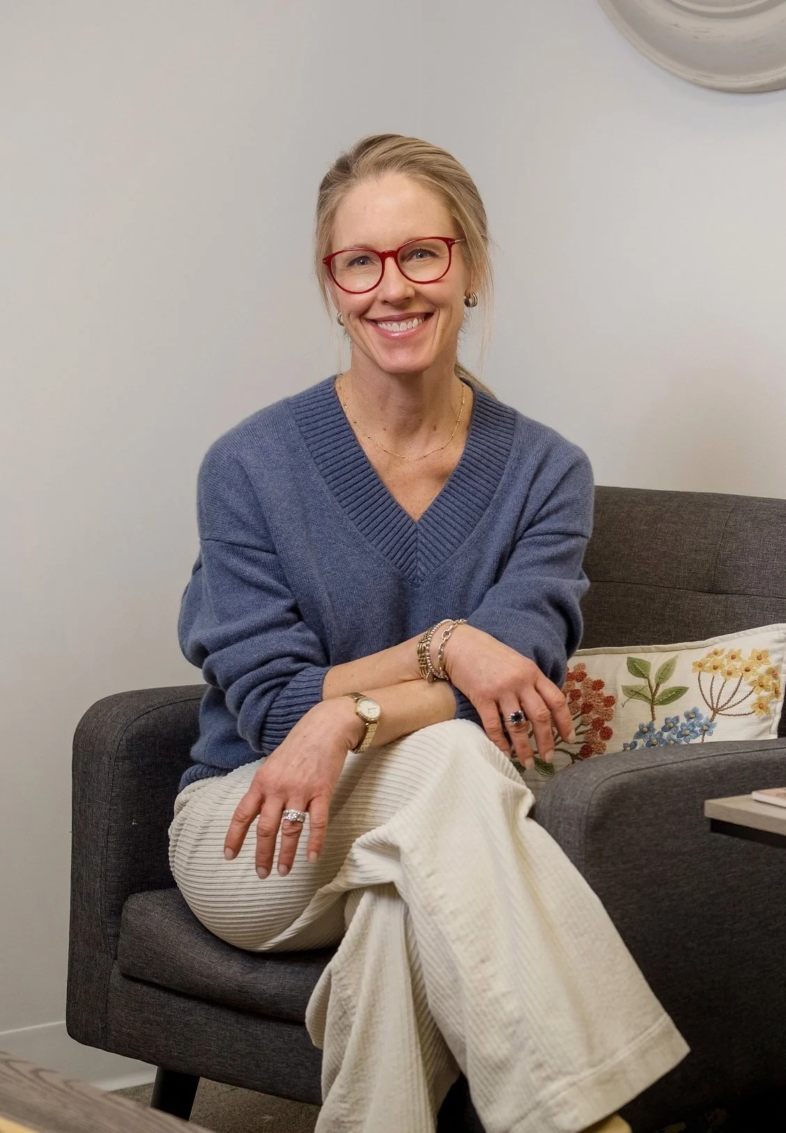 Angie Martin, Oak Park IL therapist, wearing red glasses, a blue sweater, and cream-colored pants sitting on a dark gray couch with her legs crossed, smiling at the camera.