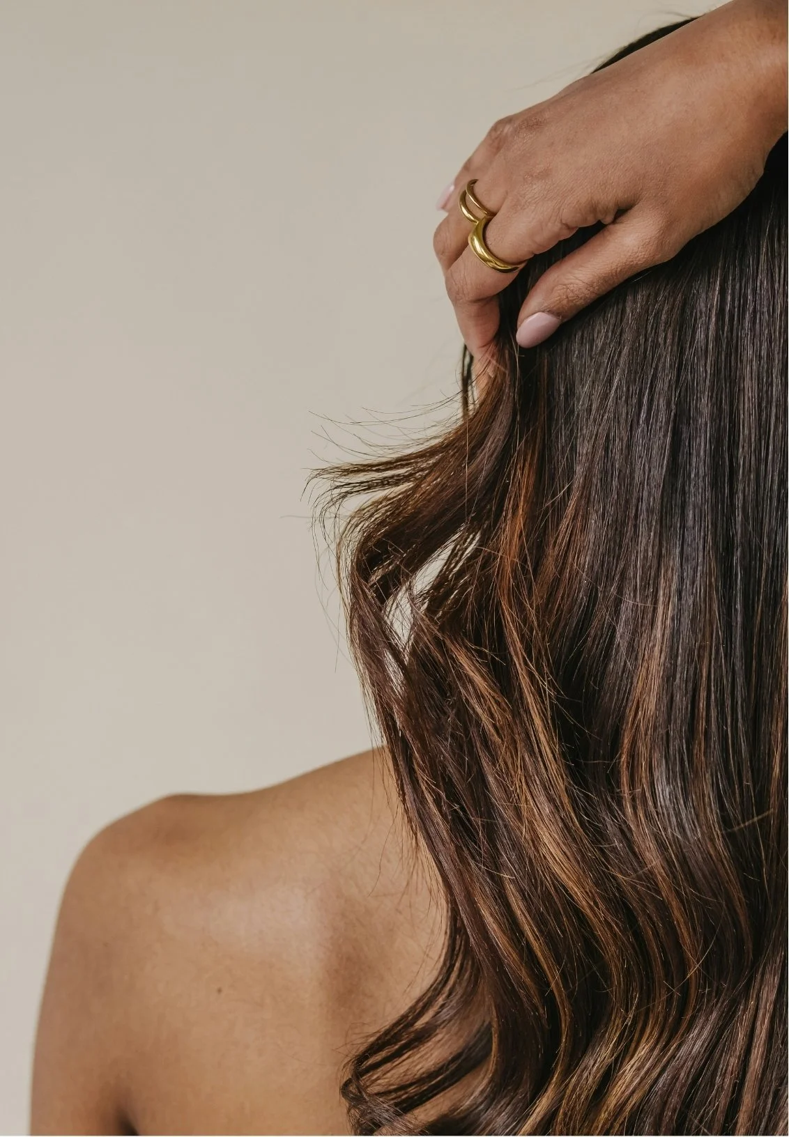 A person with wavy brown hair touching their head with their hand, wearing gold rings on fingers, and a bare shoulder visible against a plain background.