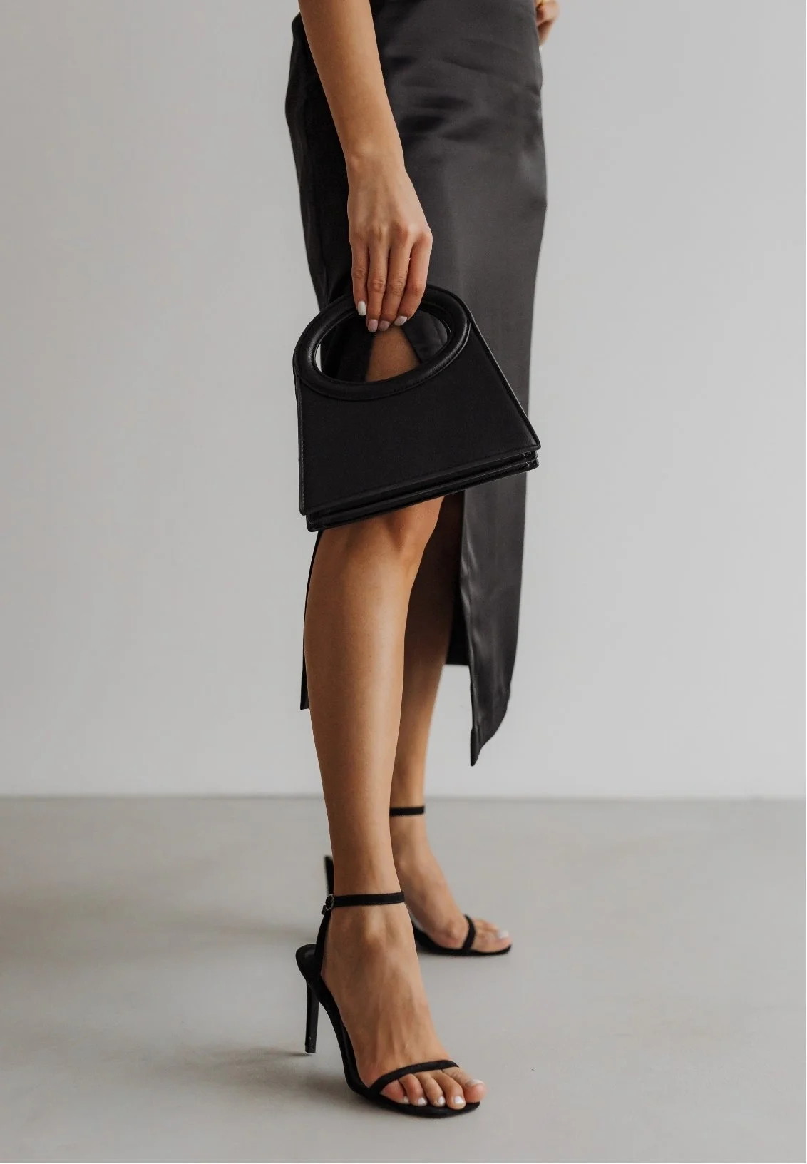 A woman dressed in a black dress, holding a black handbag, and wearing black high-heeled sandals.