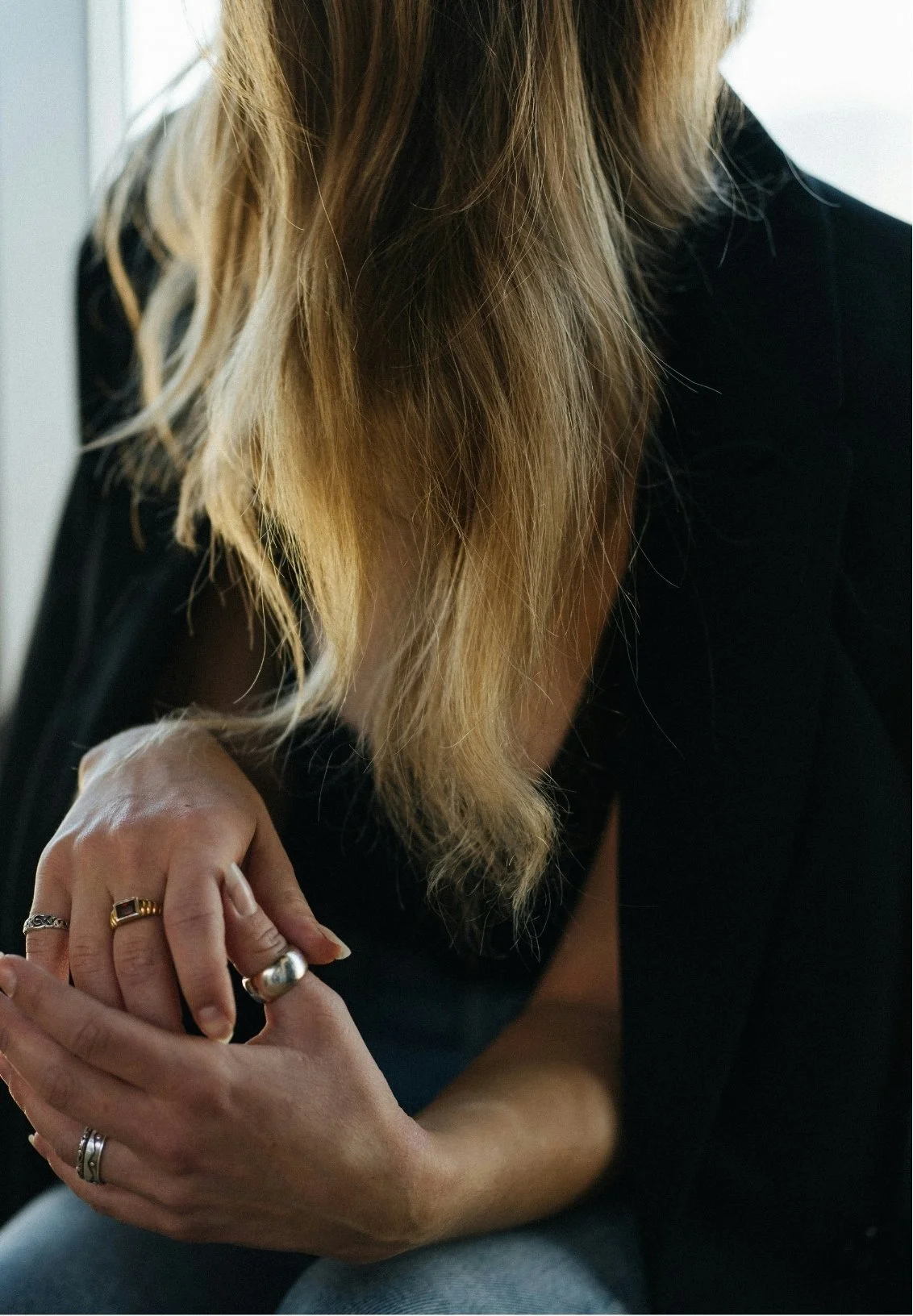 A person with long blonde hair wearing a black top, resting their arm on their knee, showing multiple rings on their fingers.